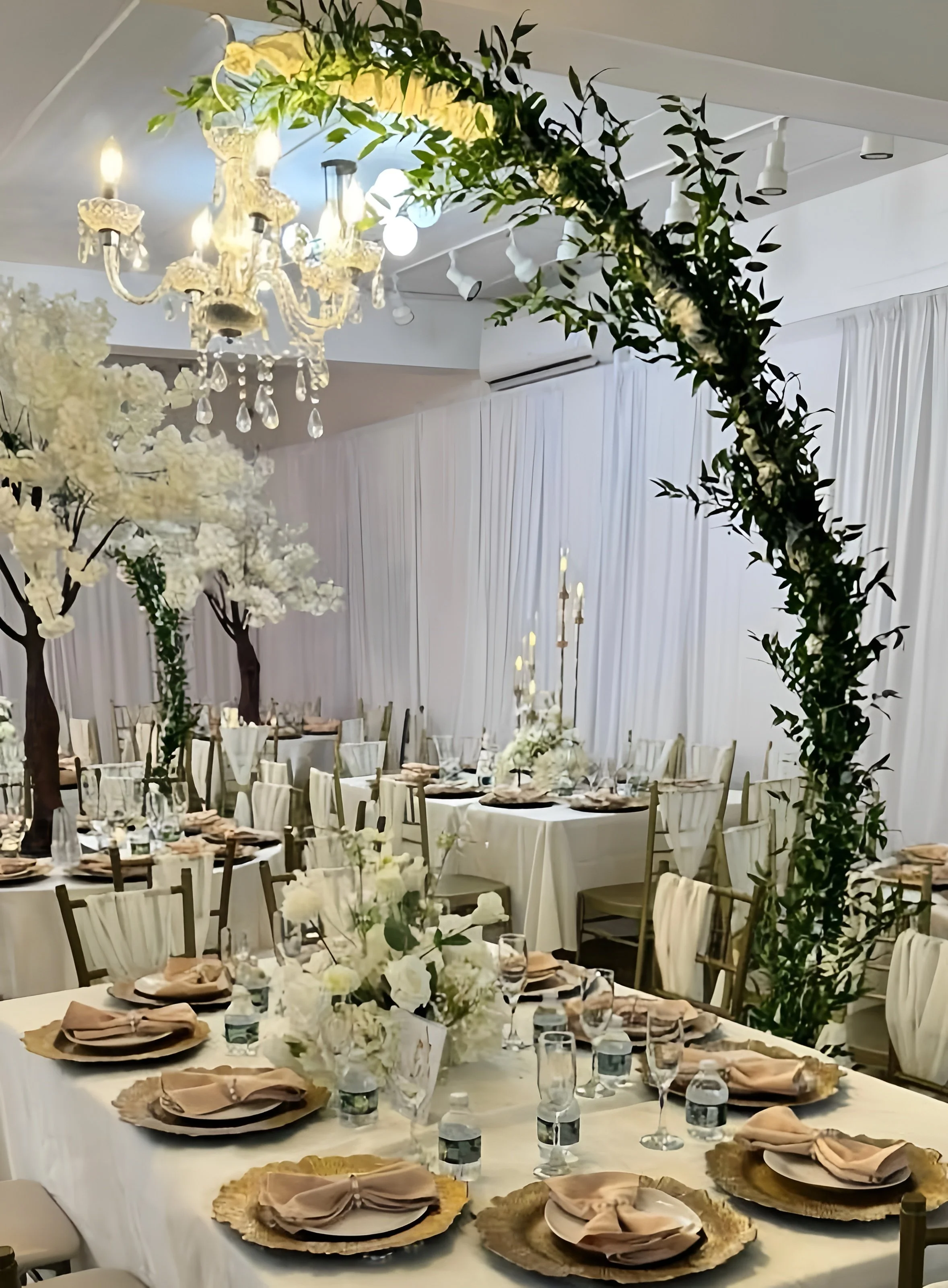 Elegant indoor wedding reception with white draped walls, decorated tables with gold chargers and napkins, tall floral centerpieces, green foliage, and a chandelier hanging from the ceiling.
