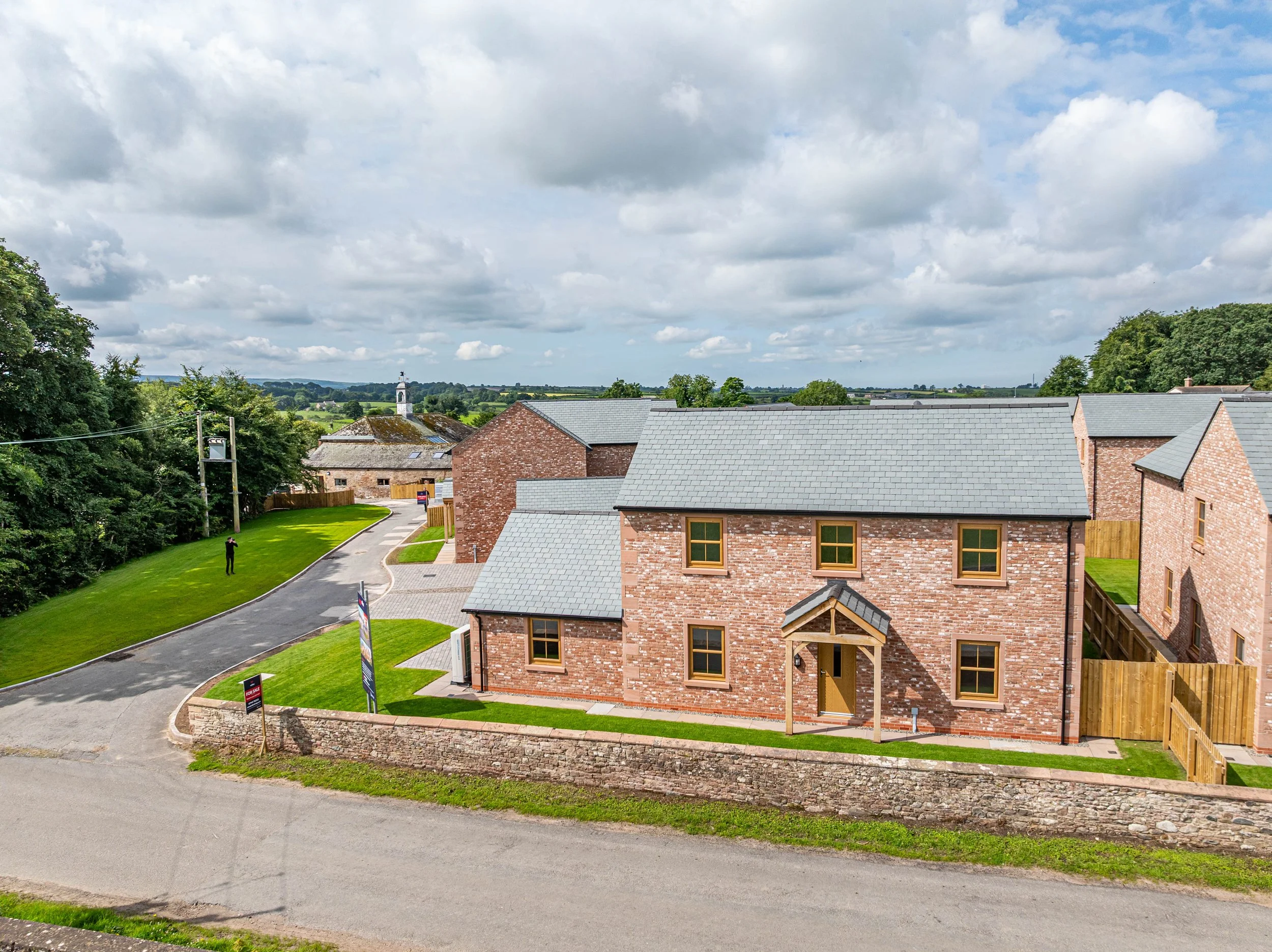 A modern brick house with a gray shingle roof and a small front porch in a suburban neighborhood. The front yard is green with a low stone wall along the sidewalk. A winding road runs past the house, with a person walking on the grass and a few trees