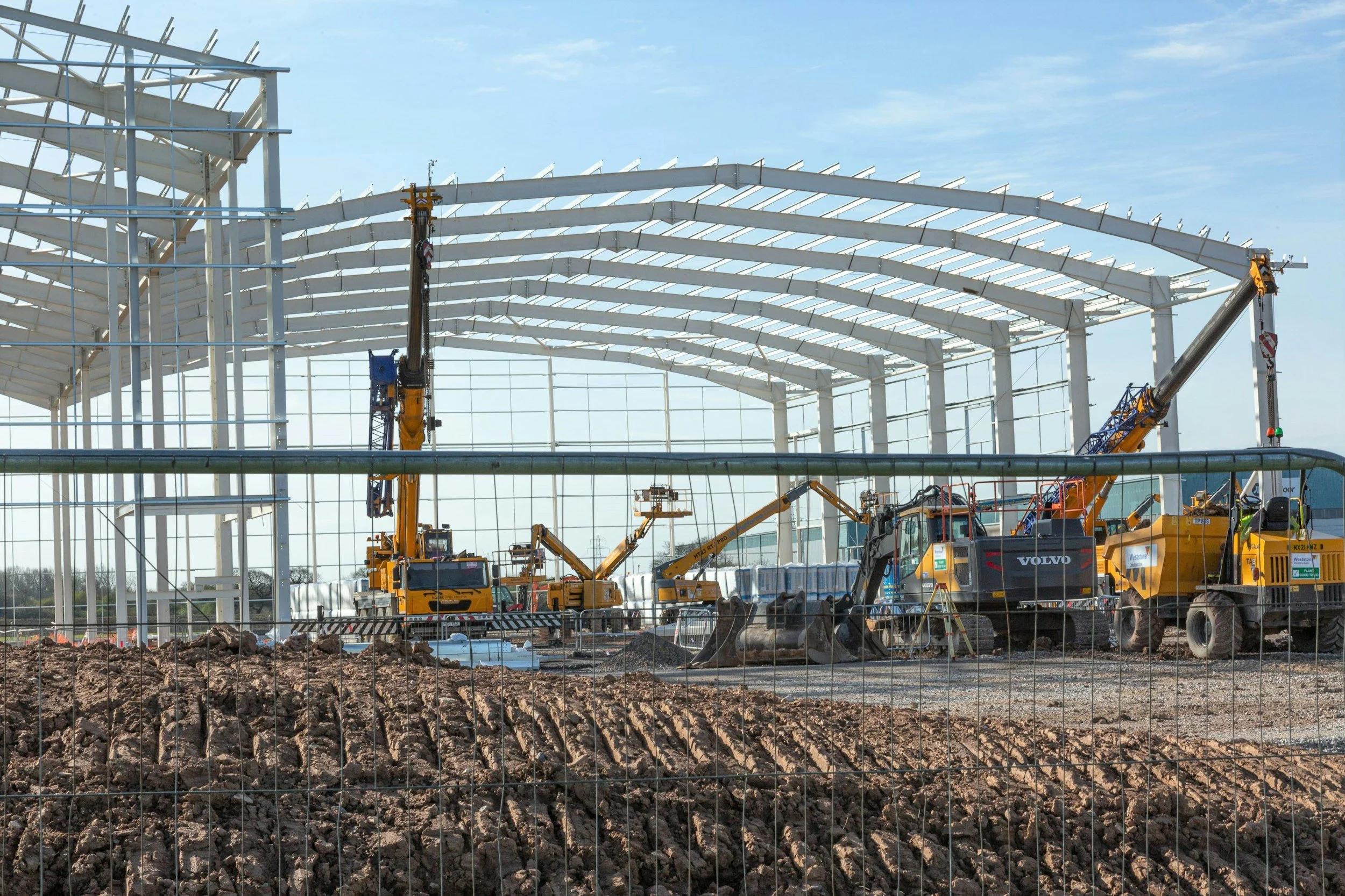 Construction site with a large structure in progress, multiple yellow cranes, and machinery working on the foundation, with a dirt area in the foreground and a partly cloudy sky.