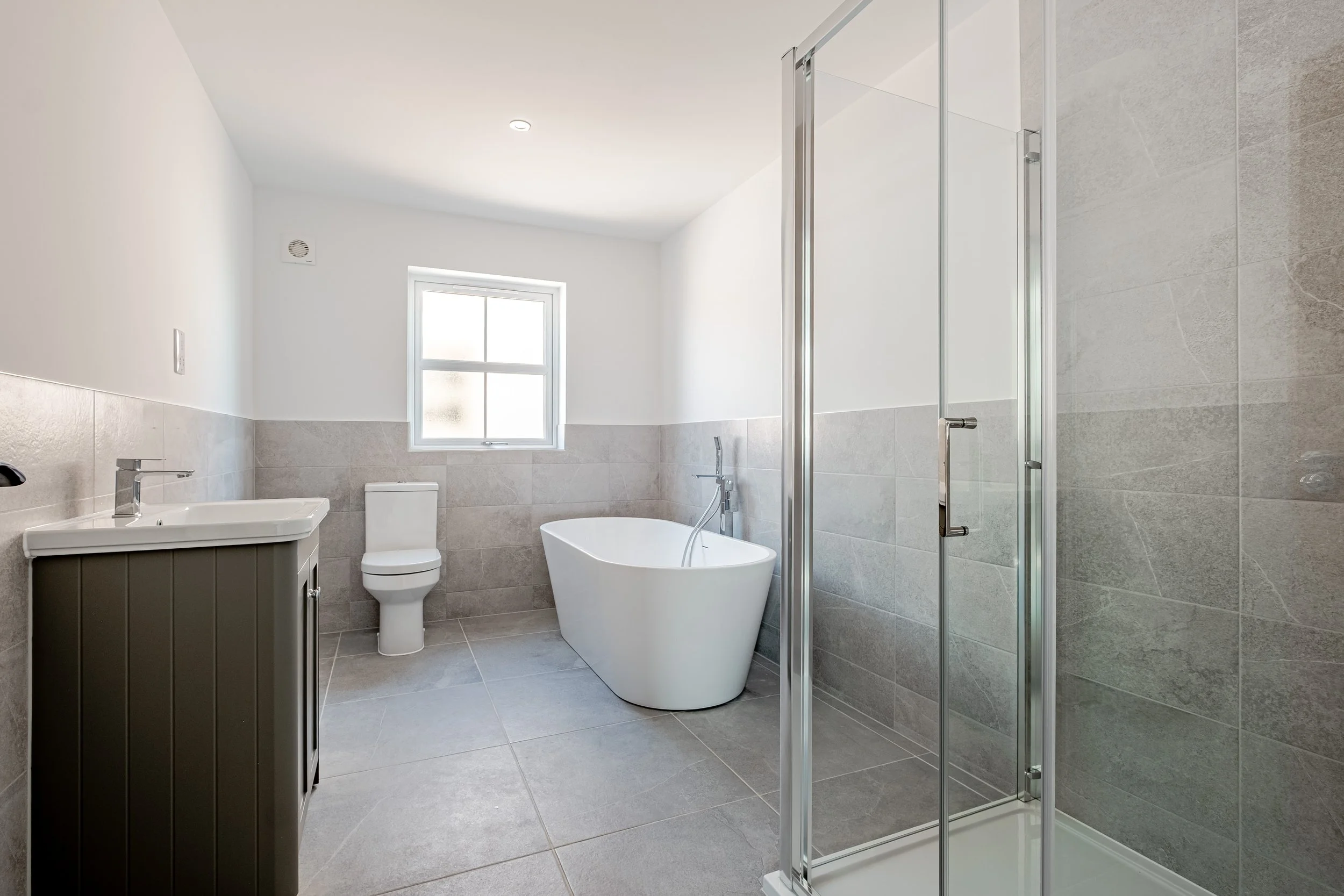 Modern bathroom with gray tiled floor and walls, a white bathtub, a toilet, a gray vanity with a sink, and a glass shower enclosure. A window allows natural light to enter.
