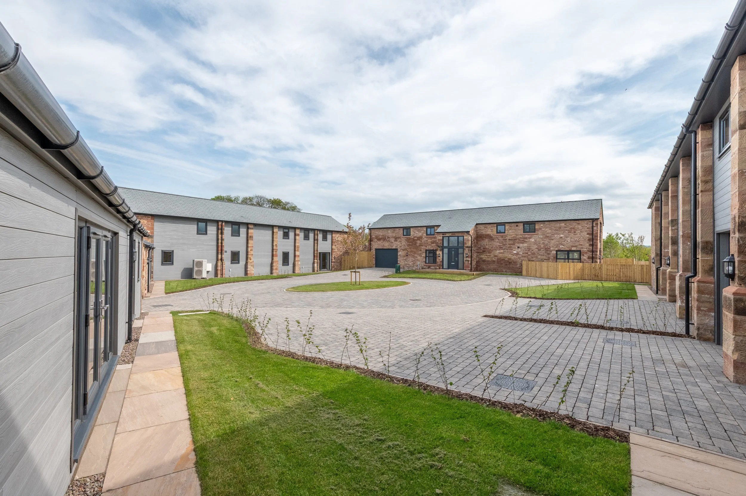 View of a residential courtyard with brick and gray buildings, paved walkways, small patches of grass, and a blue sky with clouds.