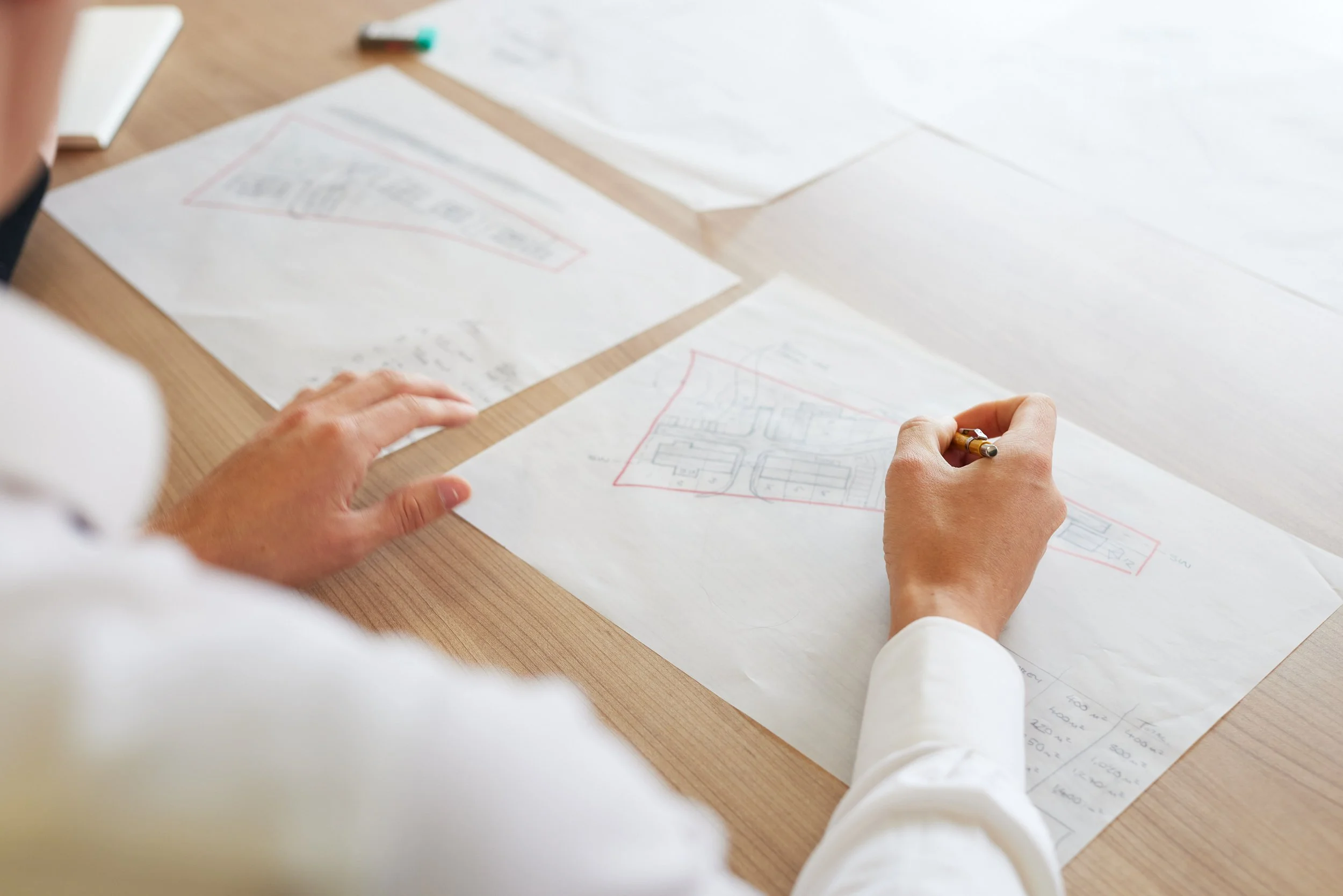 Person sketching a building floor plan on a large sheet of paper on a wooden table, with additional sketches and notes around.