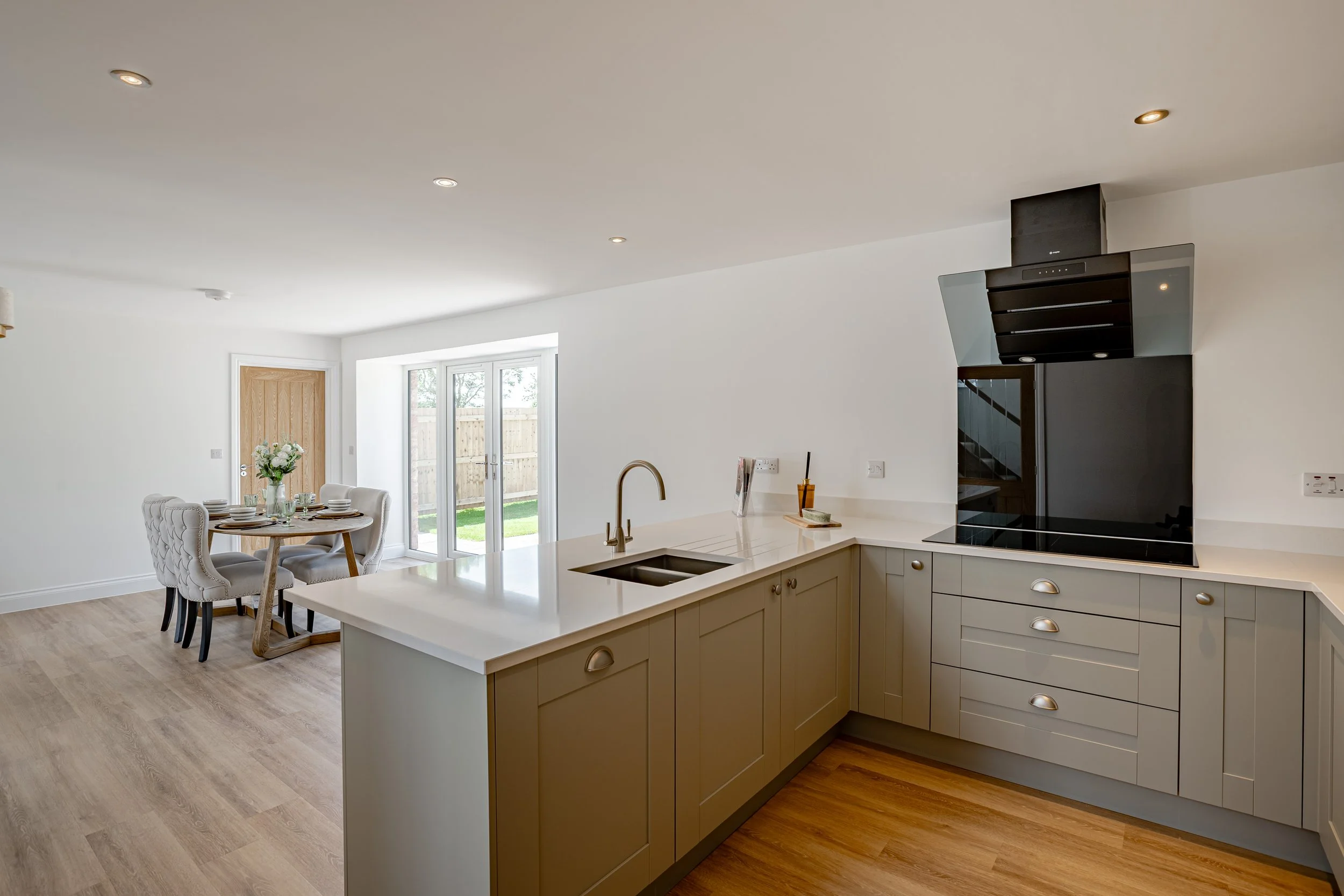 Modern open-concept kitchen with gray cabinets, white countertops, and a stove with a black and silver range hood. There's a dining area with a round table, four upholstered chairs, and a vase of flowers near sliding glass doors leading to a backyard