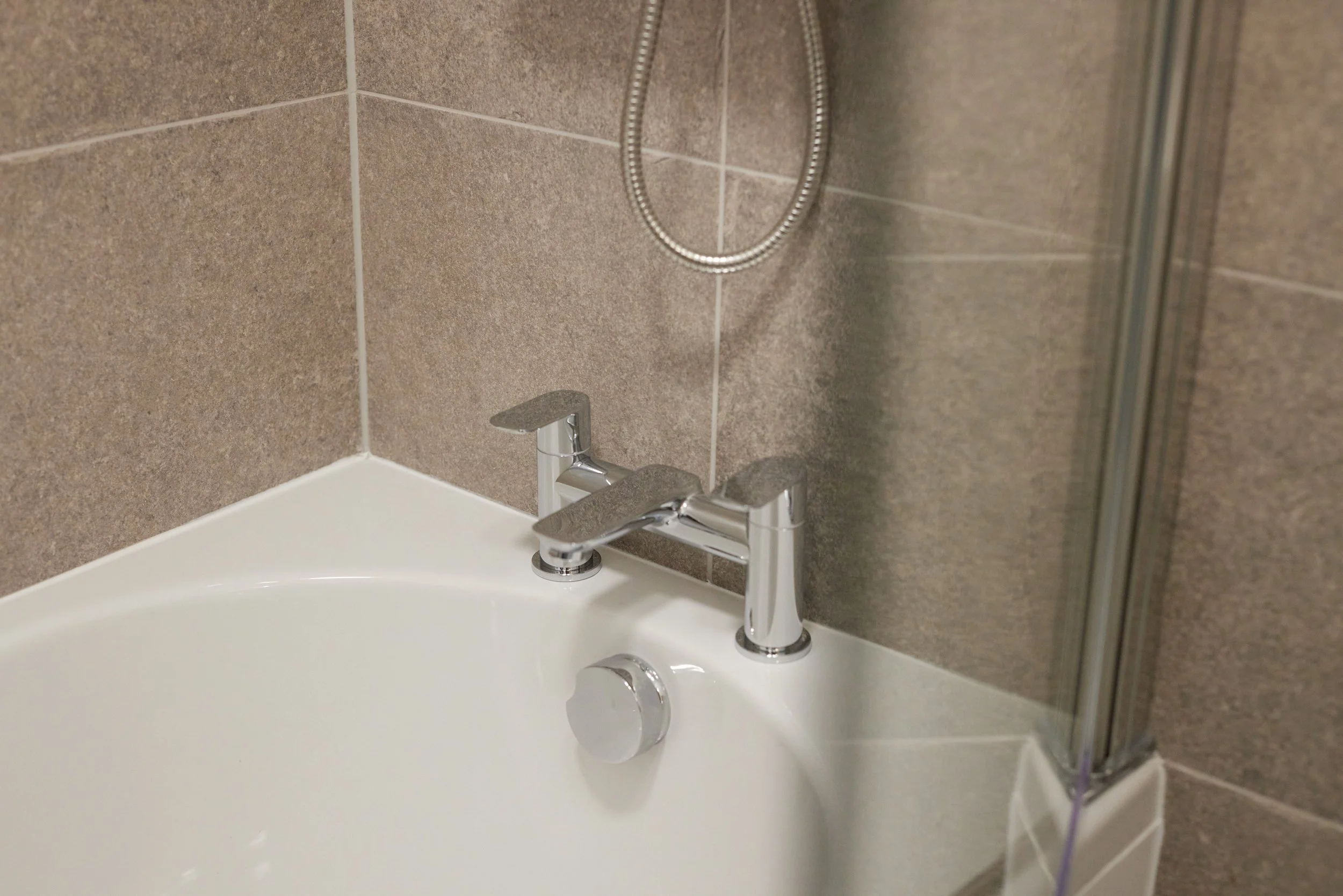 Close-up of a bathtub faucet and shower setup in a bathroom with brown tiled walls.