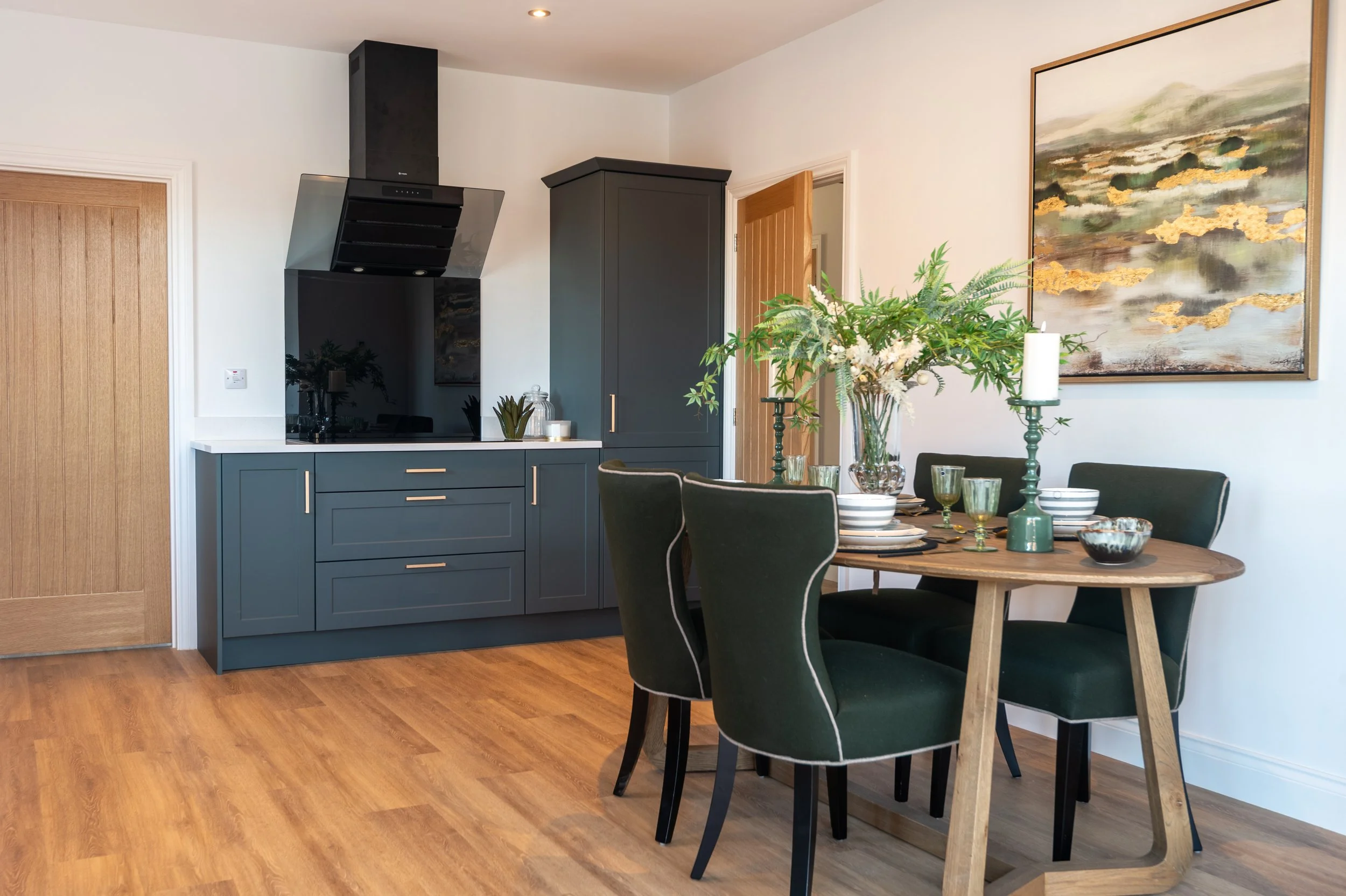 Dining room with a round wooden table, four green upholstered chairs, a large vase with white flowers, a painting on the wall, and kitchen cabinets and appliances in the background.