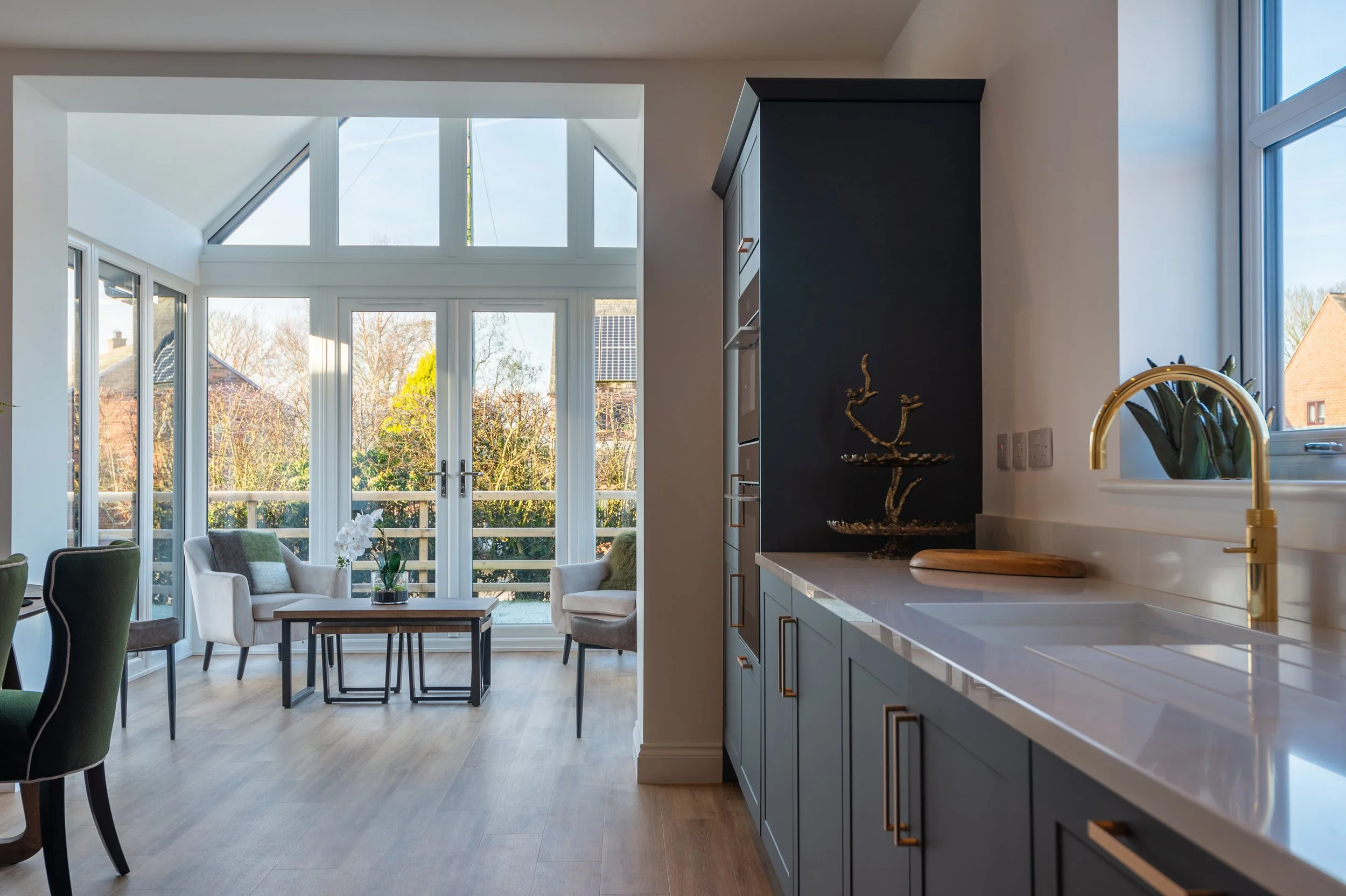 Modern kitchen with gray cabinets, white countertops, and brass faucet, adjacent to a sunlit living area with large windows and a view of trees and rooftops.