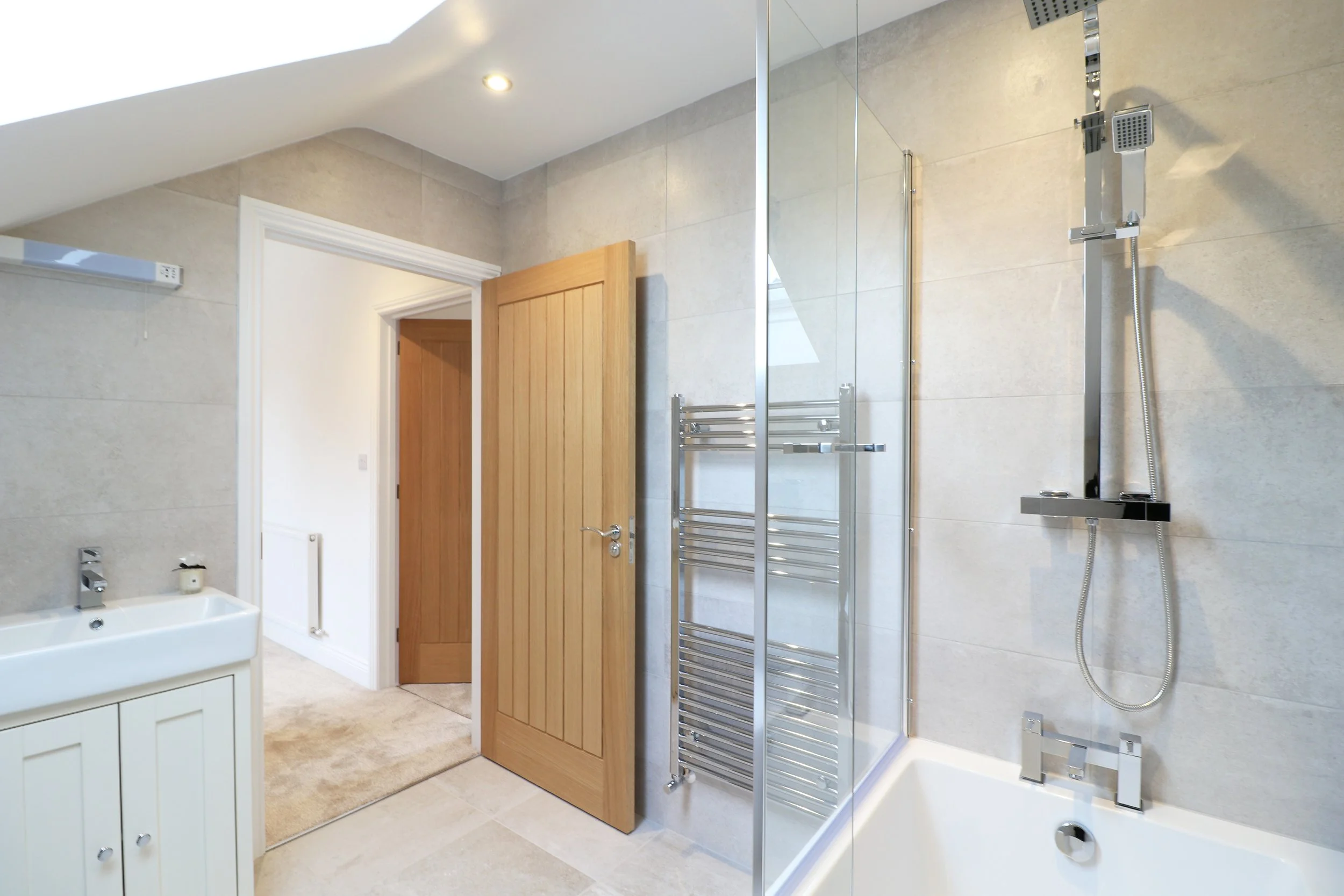 A modern bathroom with a wooden door, beige tiled walls, a white vanity with a sink, a silver towel rack, a glass-enclosed shower with a rain showerhead, and a bathtub.