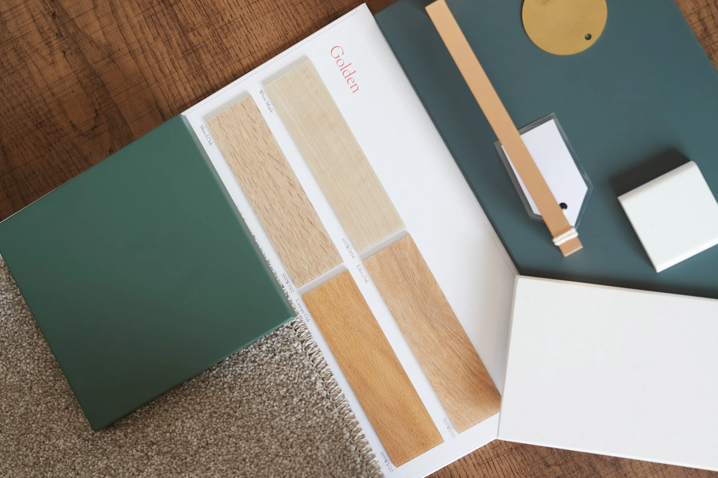 Open sample book displaying different wood finish color options, including white, yellowish beige, and darker brown shades, on a table with other sample materials nearby.