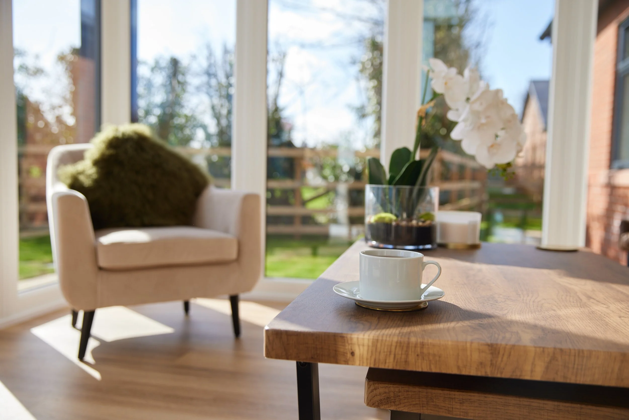 A cozy sunlit room with a wooden table, a white coffee cup on a saucer, a potted orchid, a beige armchair with a green pillow, and large windows showing a green outdoor space.