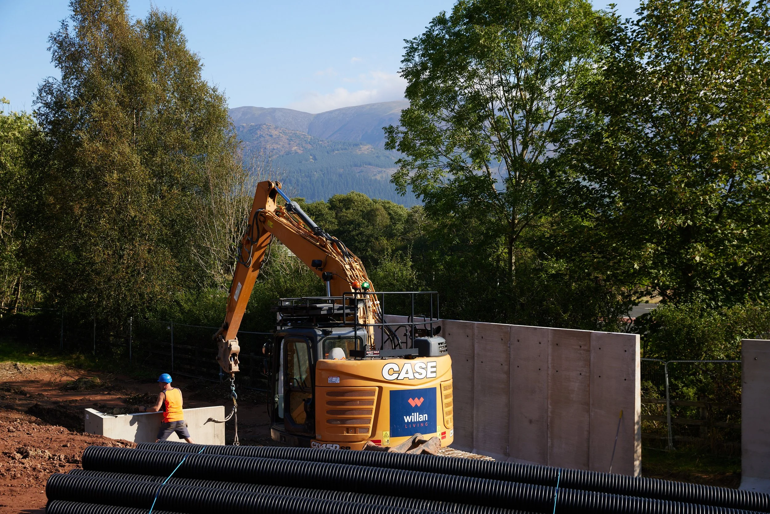Construction site with a worker in a blue helmet and orange vest using a piece of heavy machinery, with trees and mountains in the background.