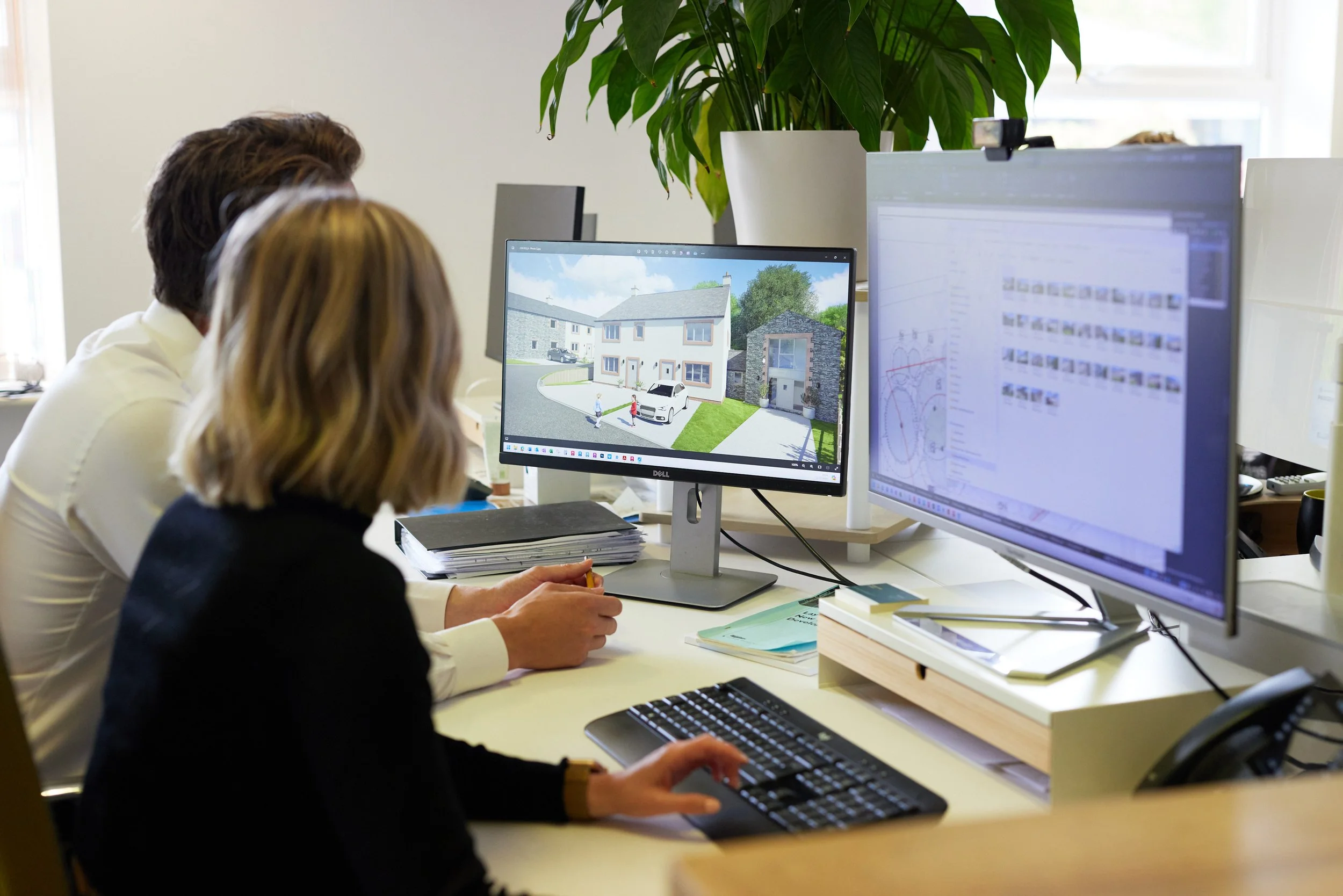 Two people working at a desk with two computer monitors, one displaying a 3D rendering of a neighborhood with houses and cars, and the other showing design and technical sketches, in an office with natural light and a large green potted plant.