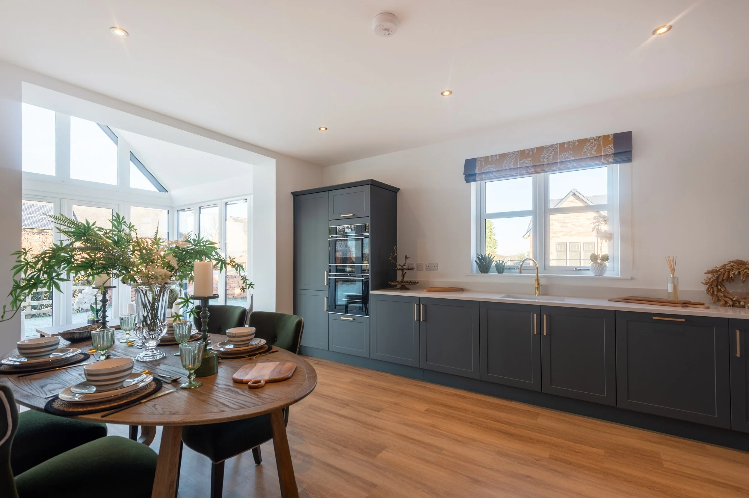 Modern kitchen with dark green cabinets, a window above the sink, a dining table set with plates and glasses, and a sunroom area with large windows and houseplants.