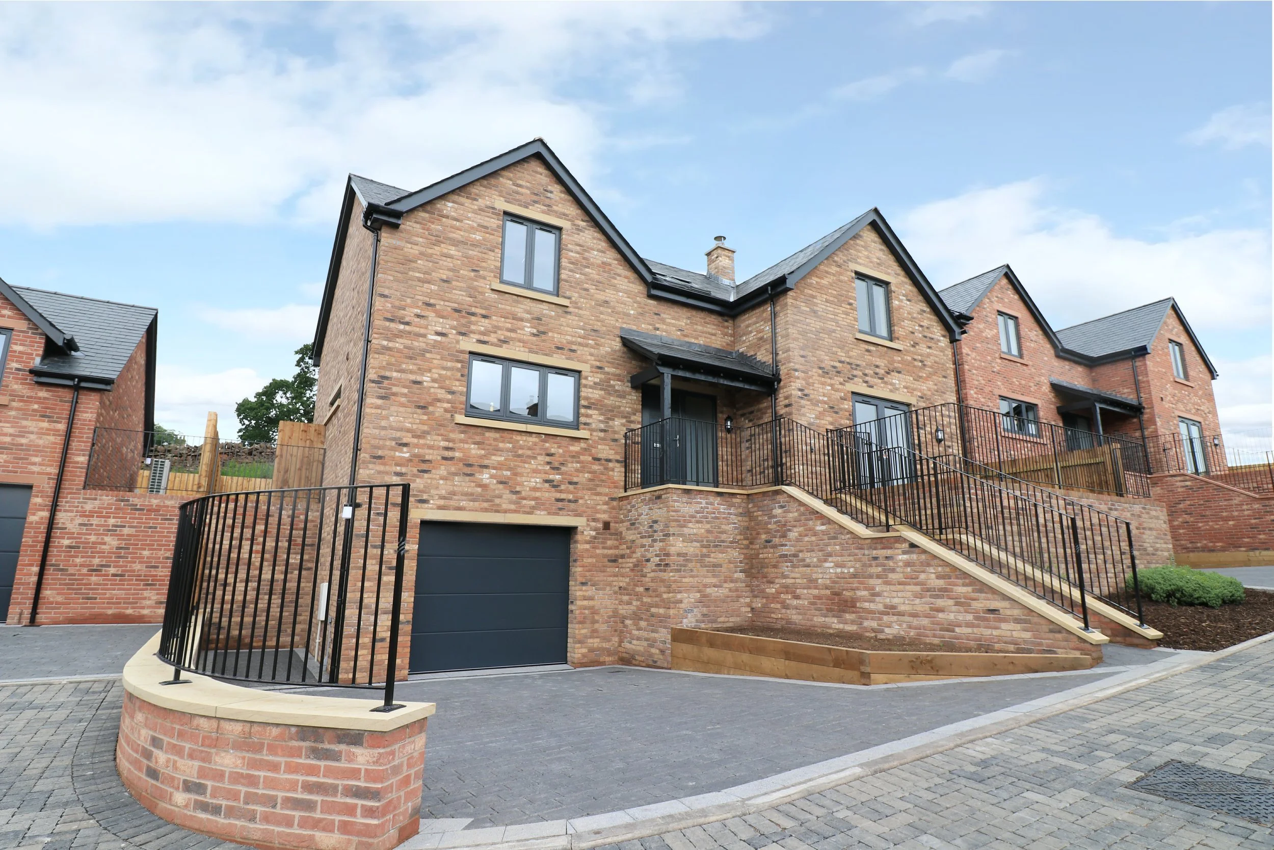 A modern brick residential building with multiple stories, black gates, and staircases leading to the front doors, surrounded by a paved driveway and landscaped areas.