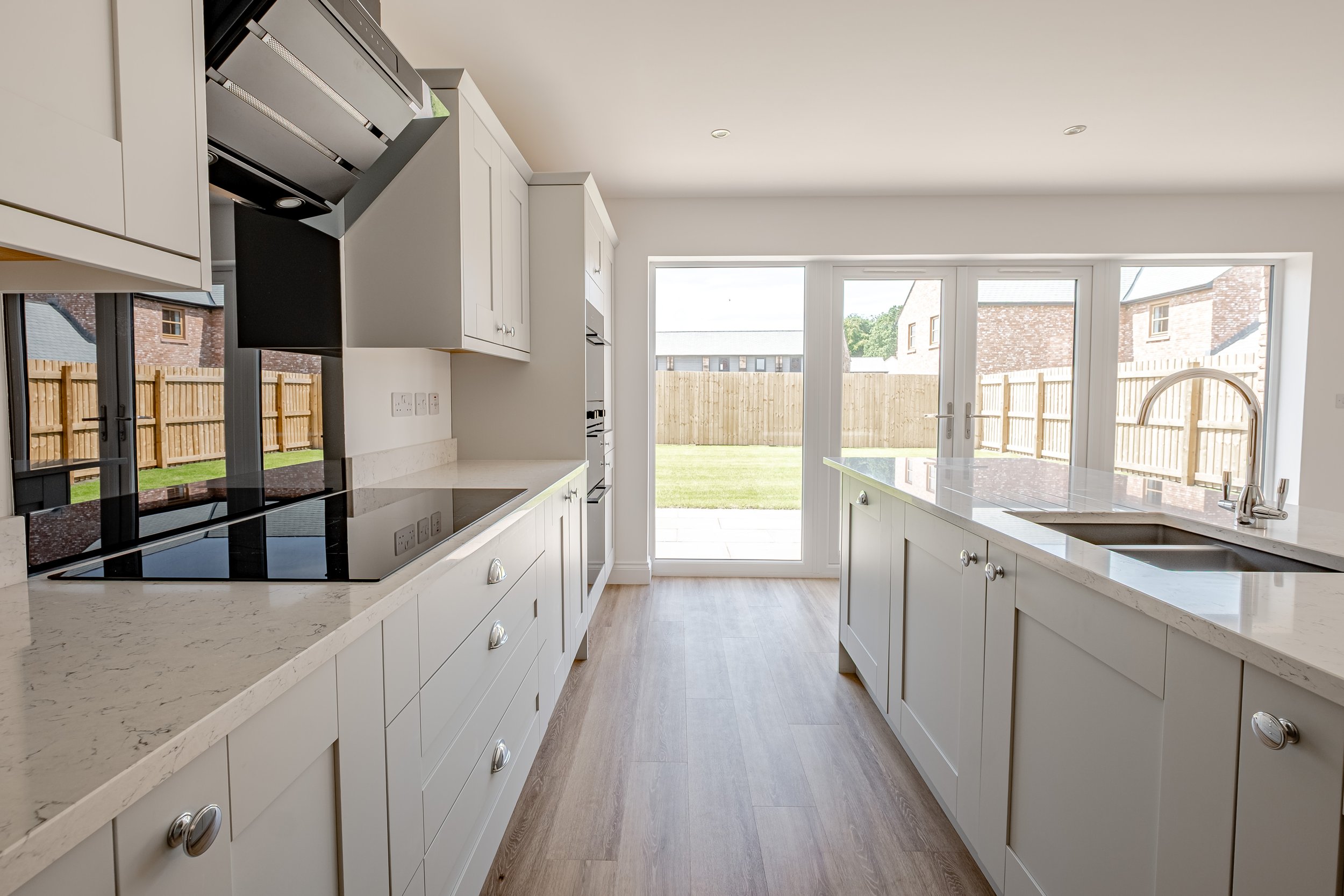 Modern kitchen with white cabinets, marble countertops, stovetop, double oven, and a large window and glass door leading to a backyard with a wooden fence.