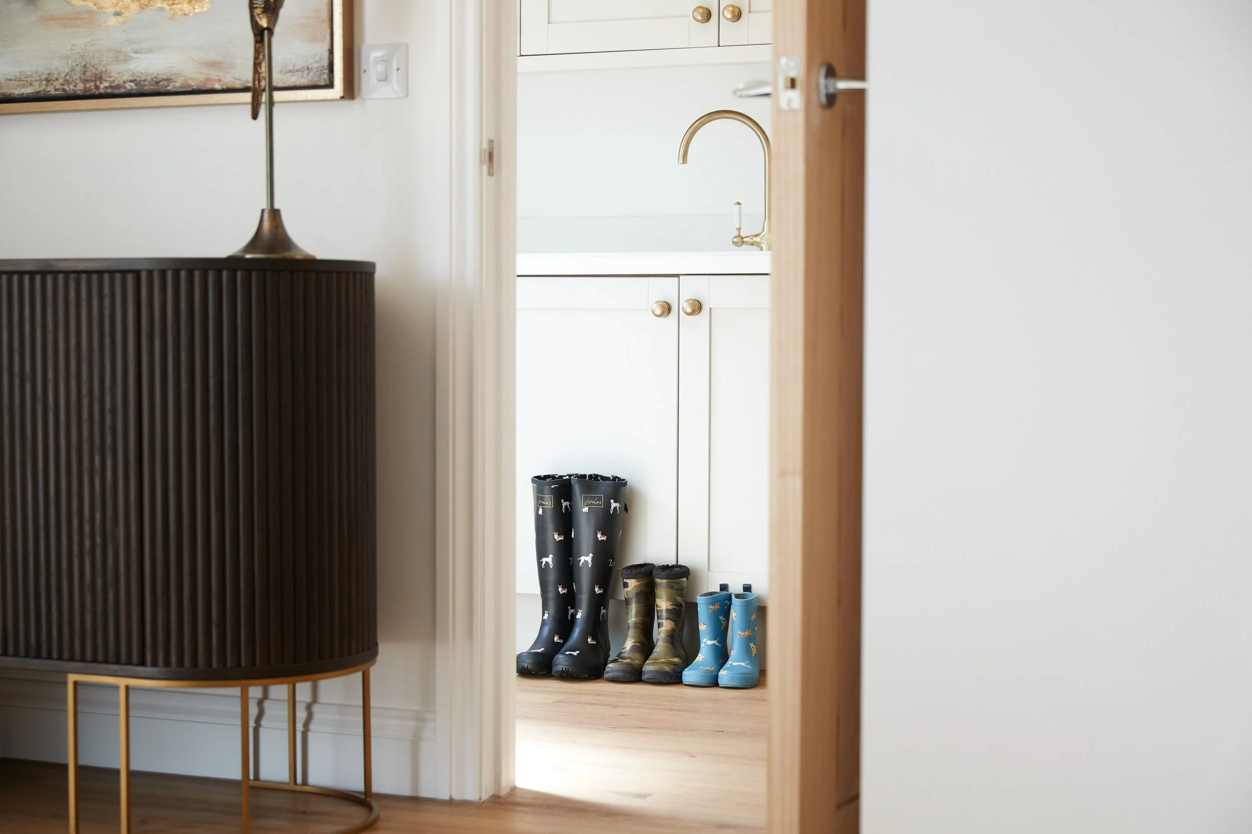 Three pairs of rain boots in black, camouflage, and blue lined up on a wooden floor in a kitchen entryway.