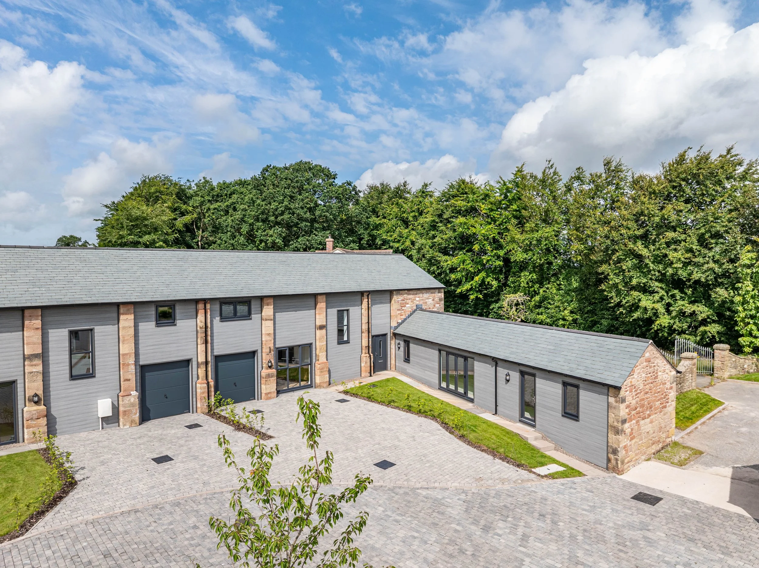A modern residential building with gray siding, stone accents, black window frames, and attached garage doors, set in a lush green area with trees and a partly cloudy sky in the background.