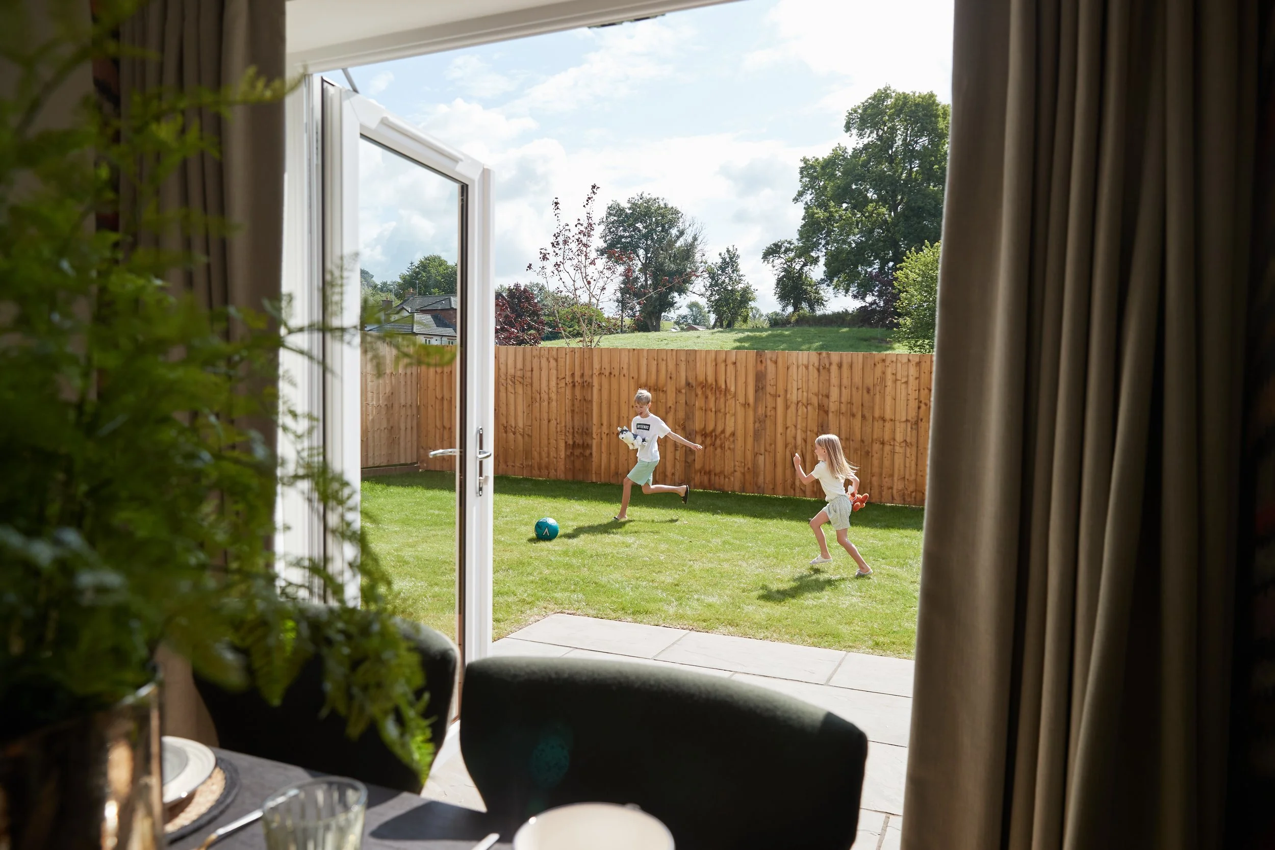 Two children playing football in a backyard seen through an open glass door from inside a house.
