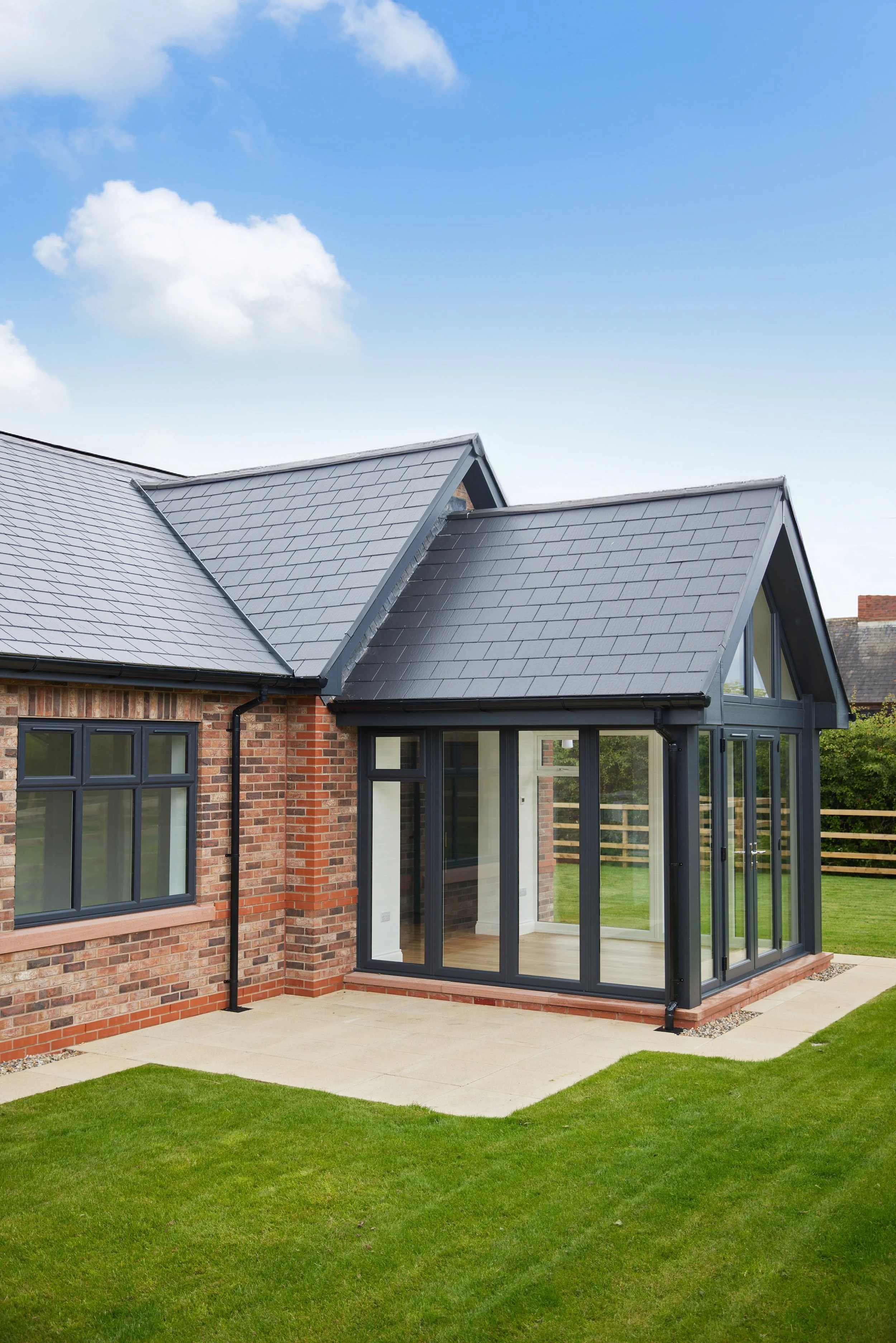 A modern brick house with a grey tiled roof and a glass sunroom extension, surrounded by a well-maintained lawn and a wooden fence, under a partly cloudy blue sky.