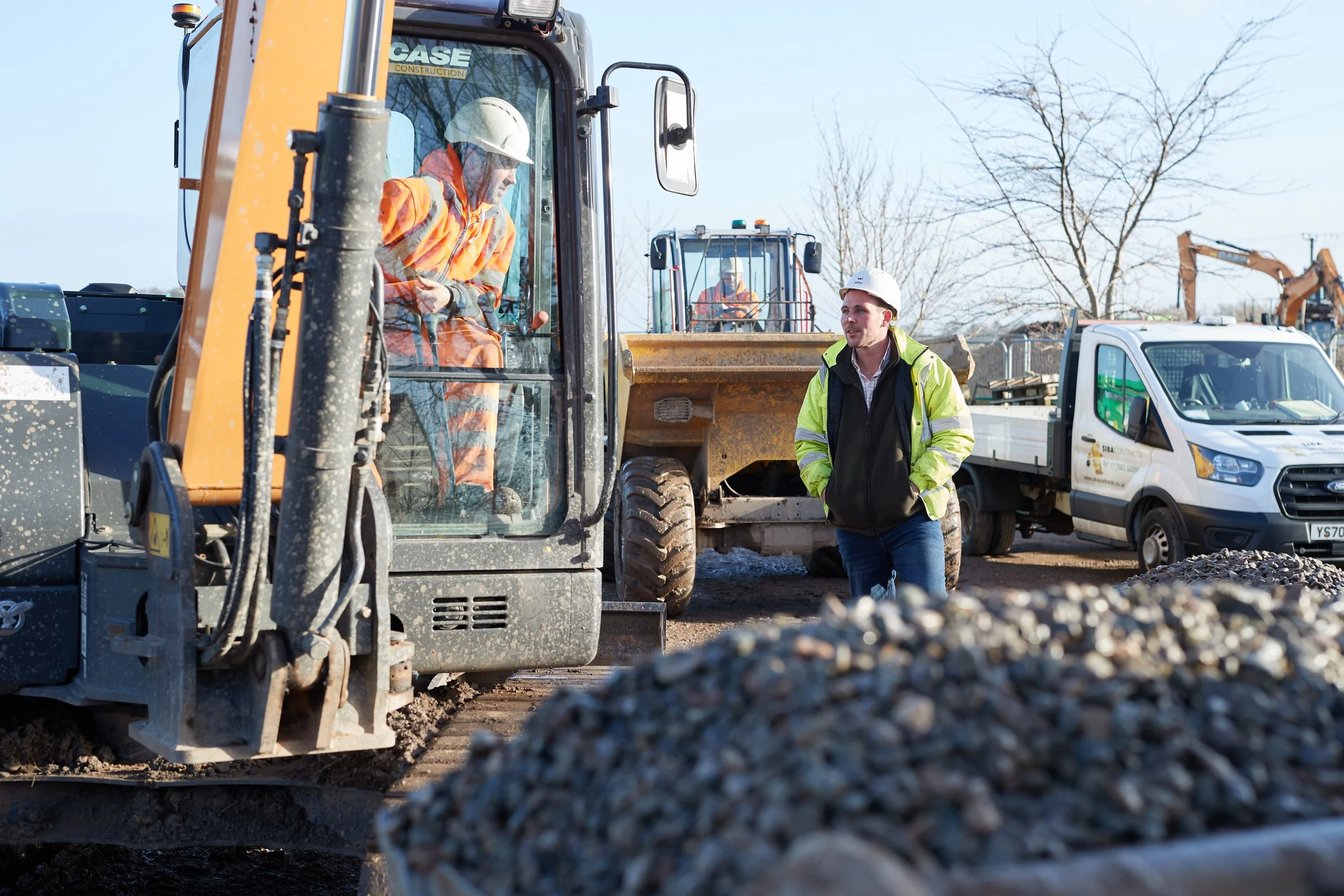 Construction site with workers, including one man in a yellow jacket and white helmet talking to a person inside a construction vehicle. Construction equipment and a white van are visible in the background, along with trees and a clear sky.