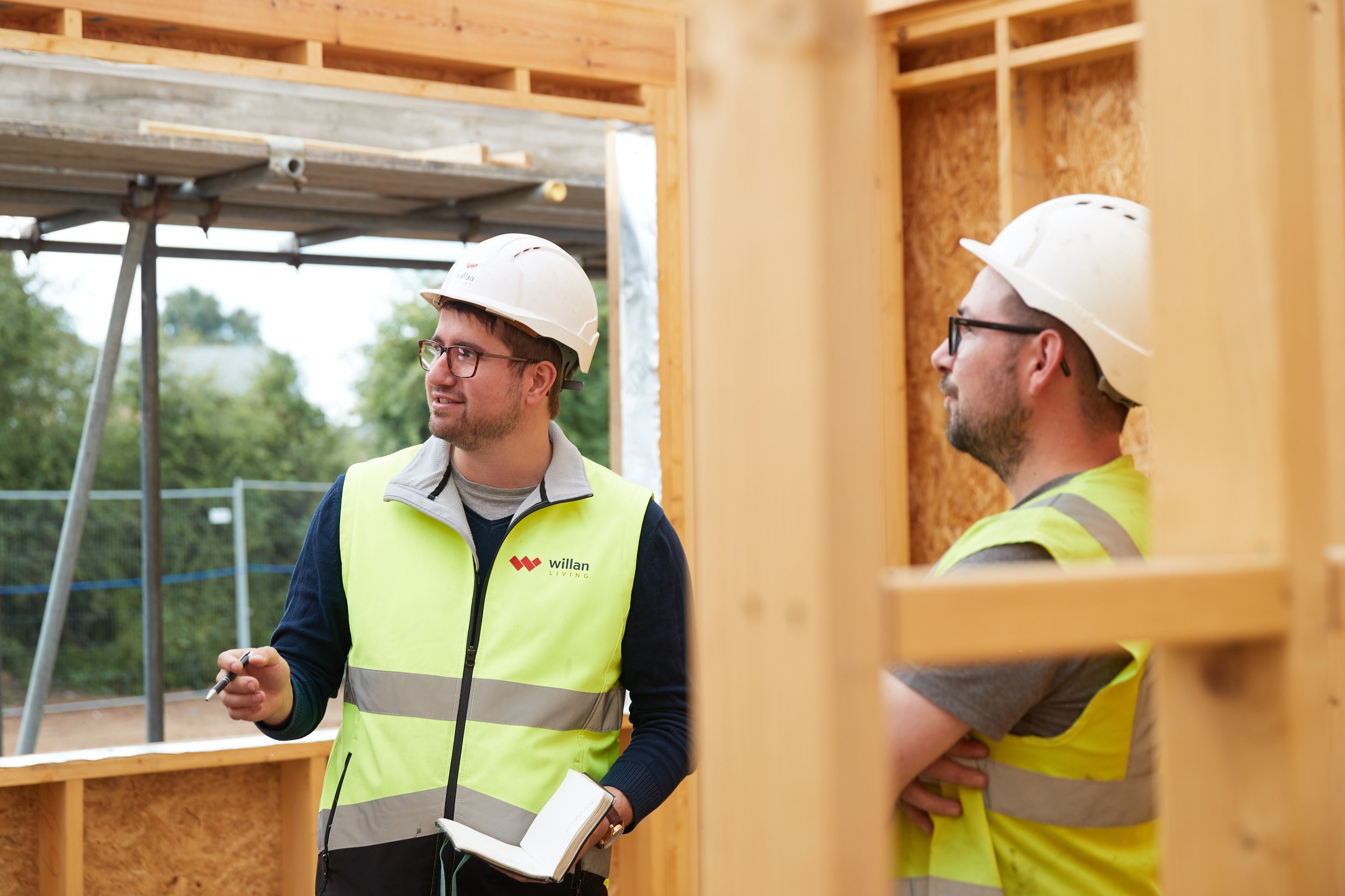 Two construction workers in safety vests and hard hats discussing on a construction site, with wooden framing and green foliage in the background.