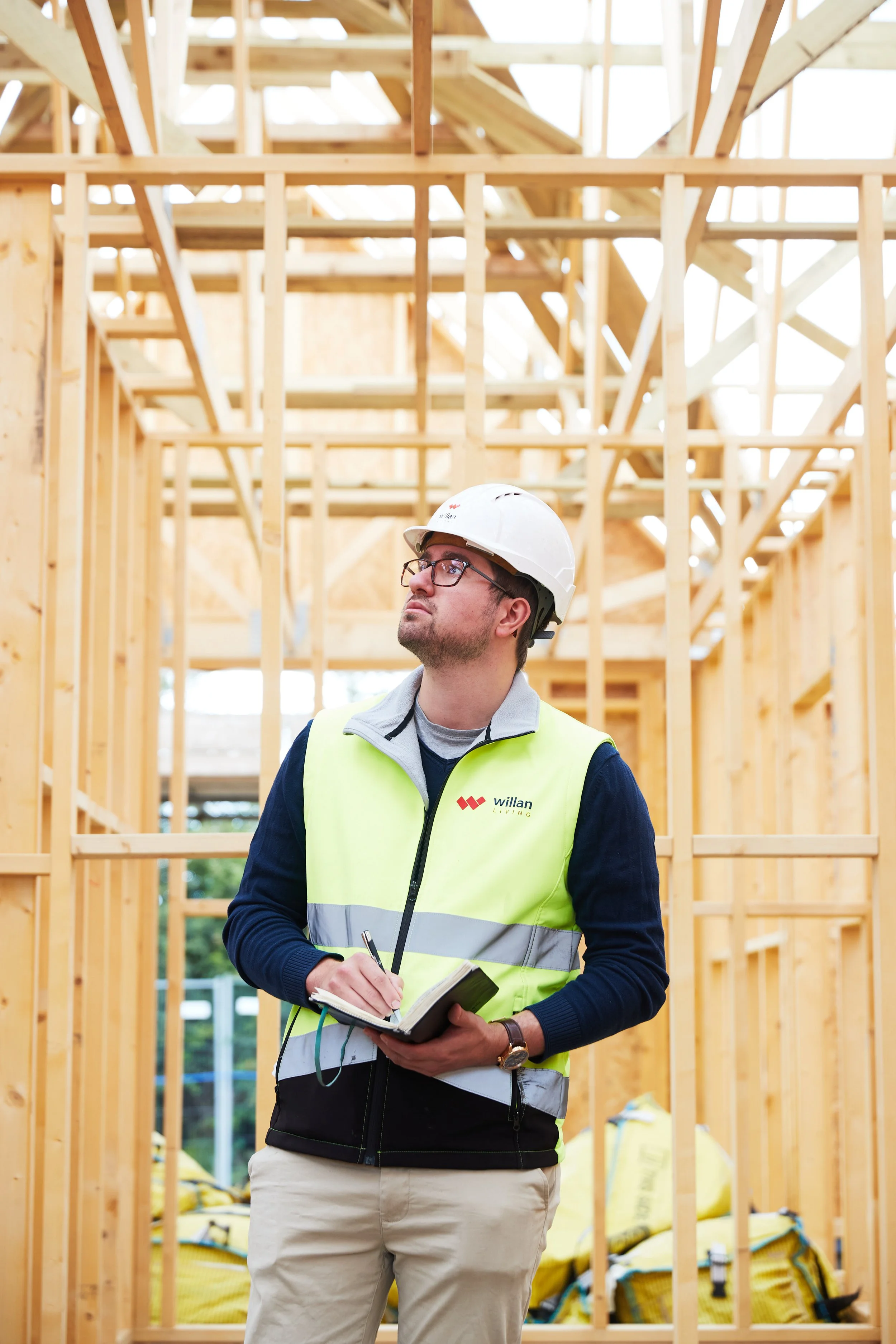 A construction worker wearing a white safety helmet, glasses, and a high-visibility vest, holding a notebook and pen, standing in front of a wooden framing of a house under construction.