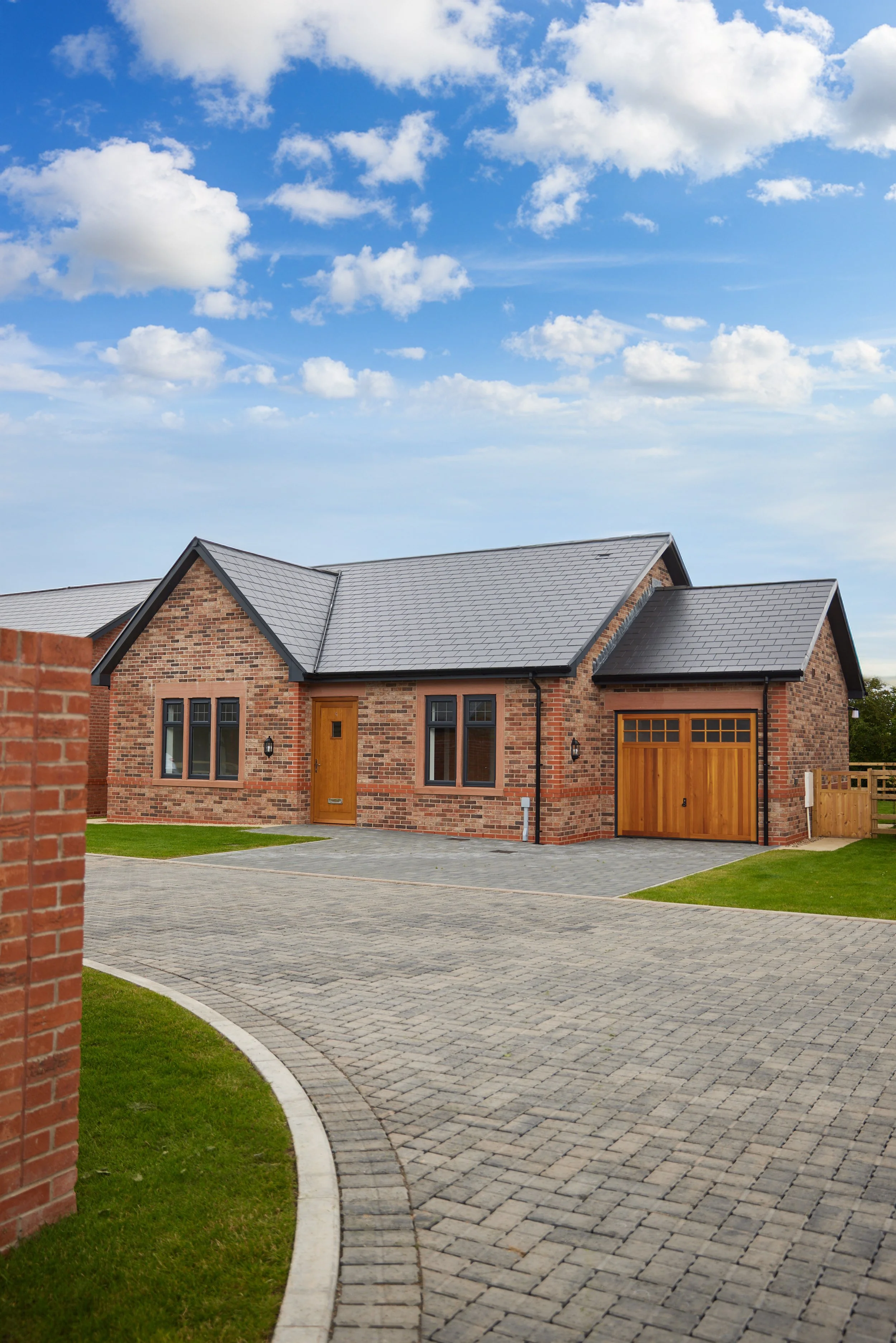 A modern brick house with a gray shingled roof, a wooden front door, and a attached garage with wooden doors. There is a curved stone driveway, green lawns, and a blue sky with clouds.