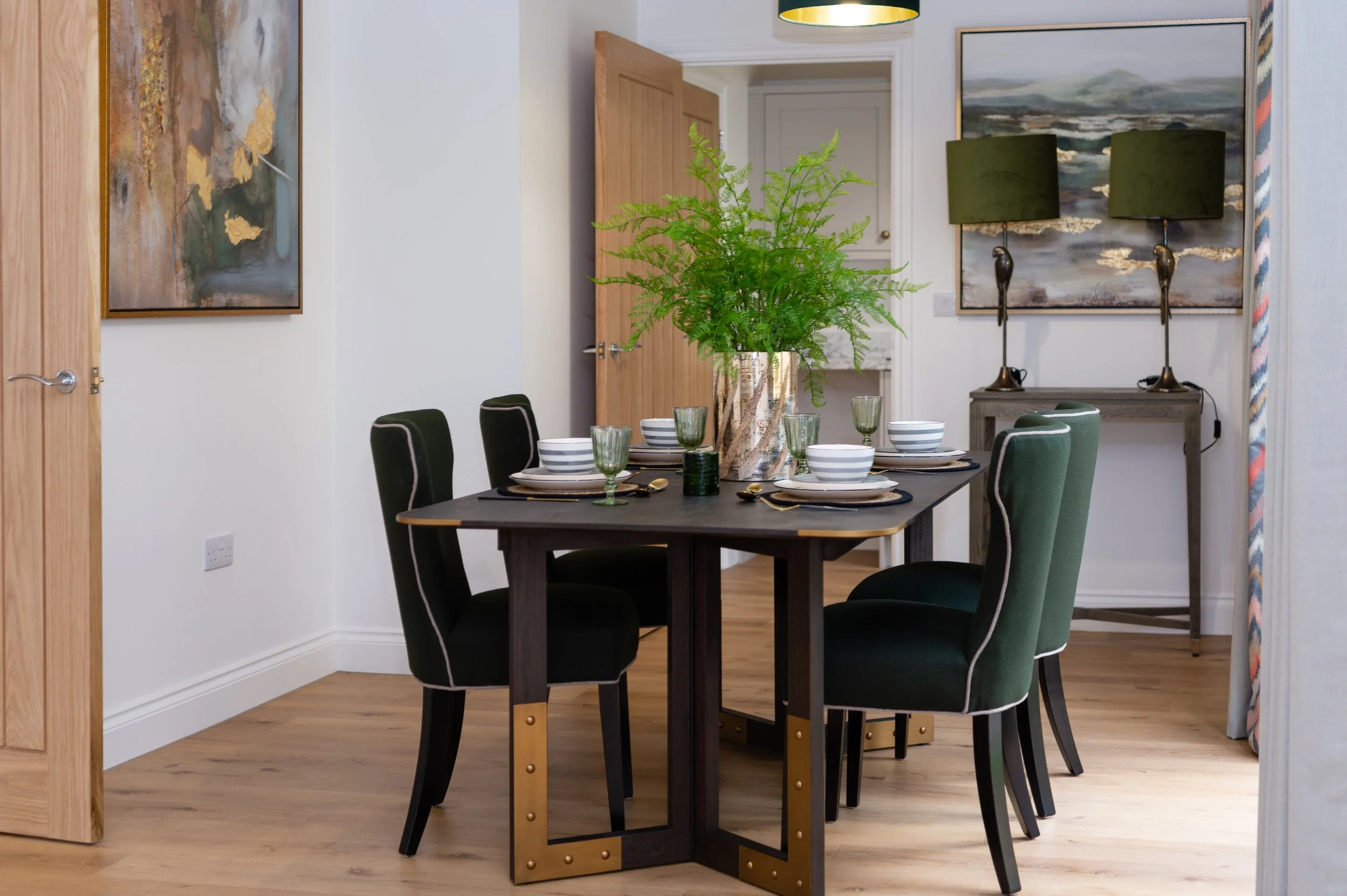 Dining room with a dark rectangular table, six green upholstered chairs, and a large leafy plant on the table. Wall art and lamps are visible in the background.