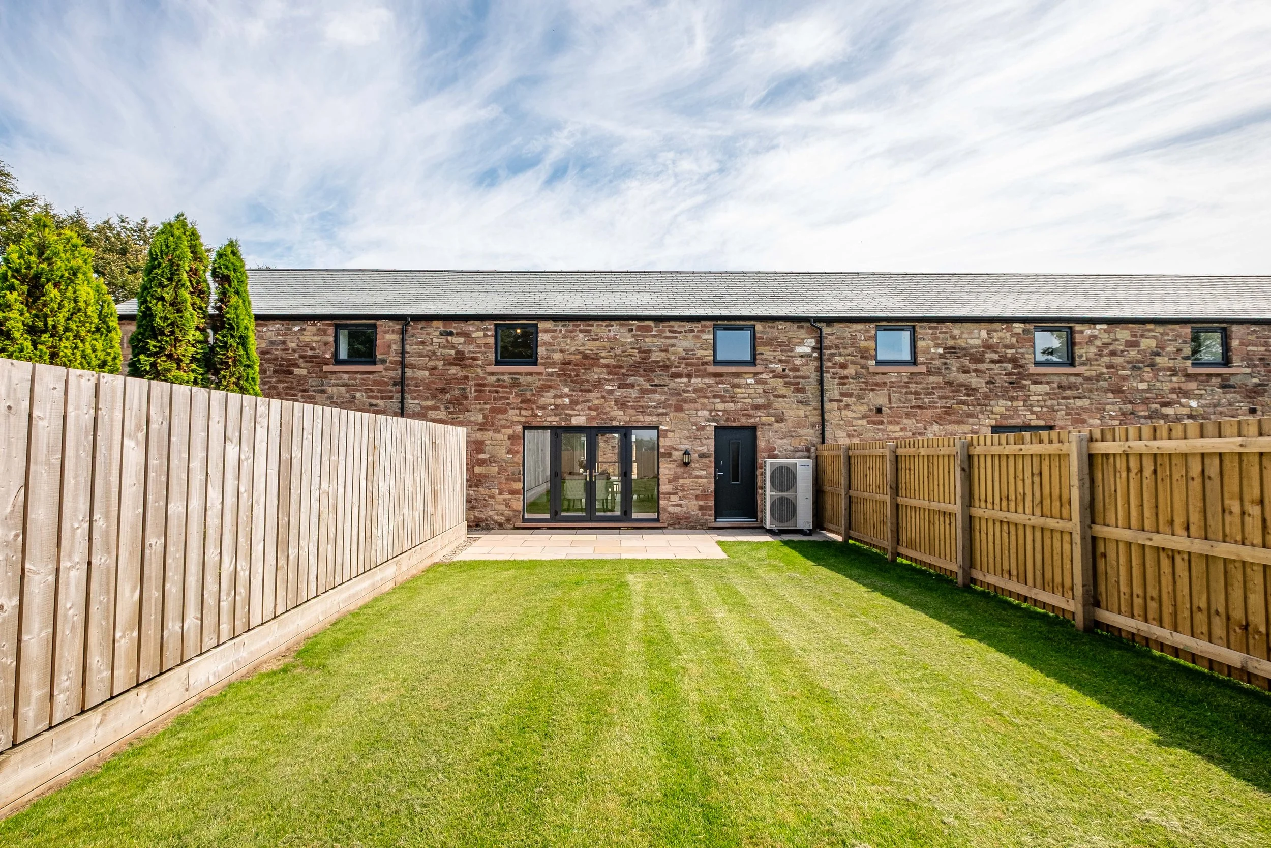 Backyard with green lawn, stone patio, wooden fencing on both sides, brick house with black-framed windows and glass doors, air conditioning unit outside, and a clear blue sky.