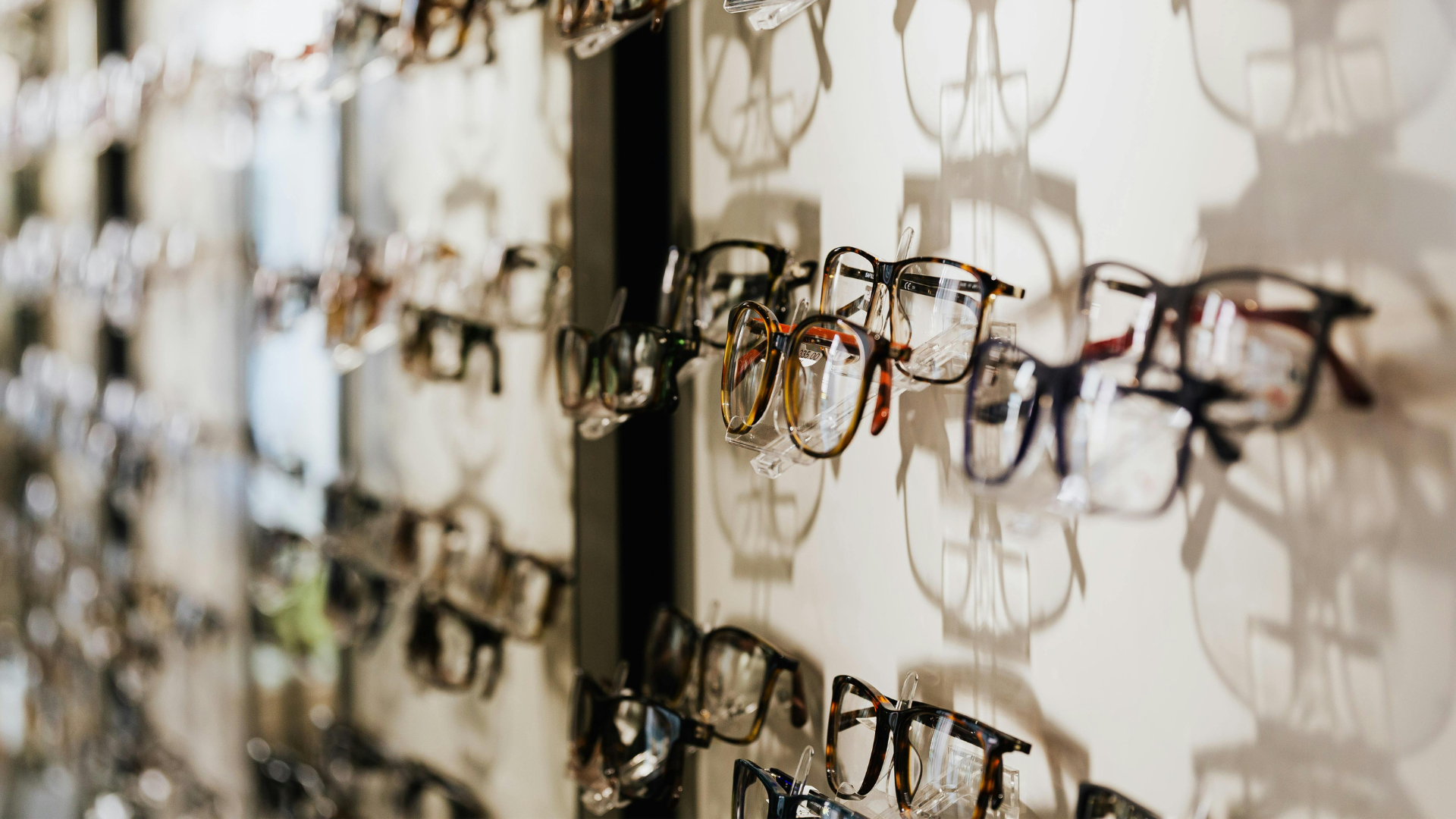A display wall with various eyeglasses and sunglasses arranged on hooks, casting shadows on the wall.