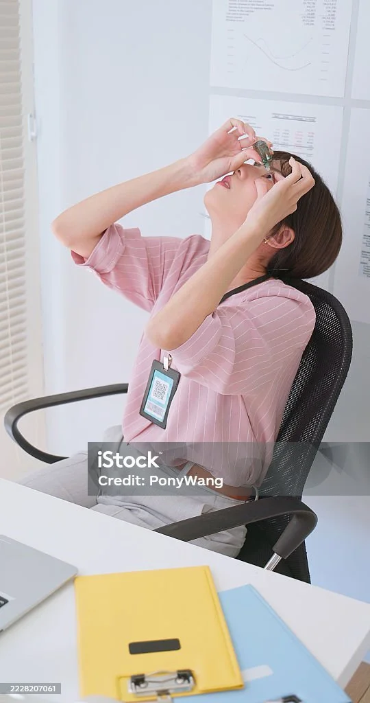 Woman wearing pink striped blouse sitting at desk with yellow and blue notebooks, looking up into her glasses.