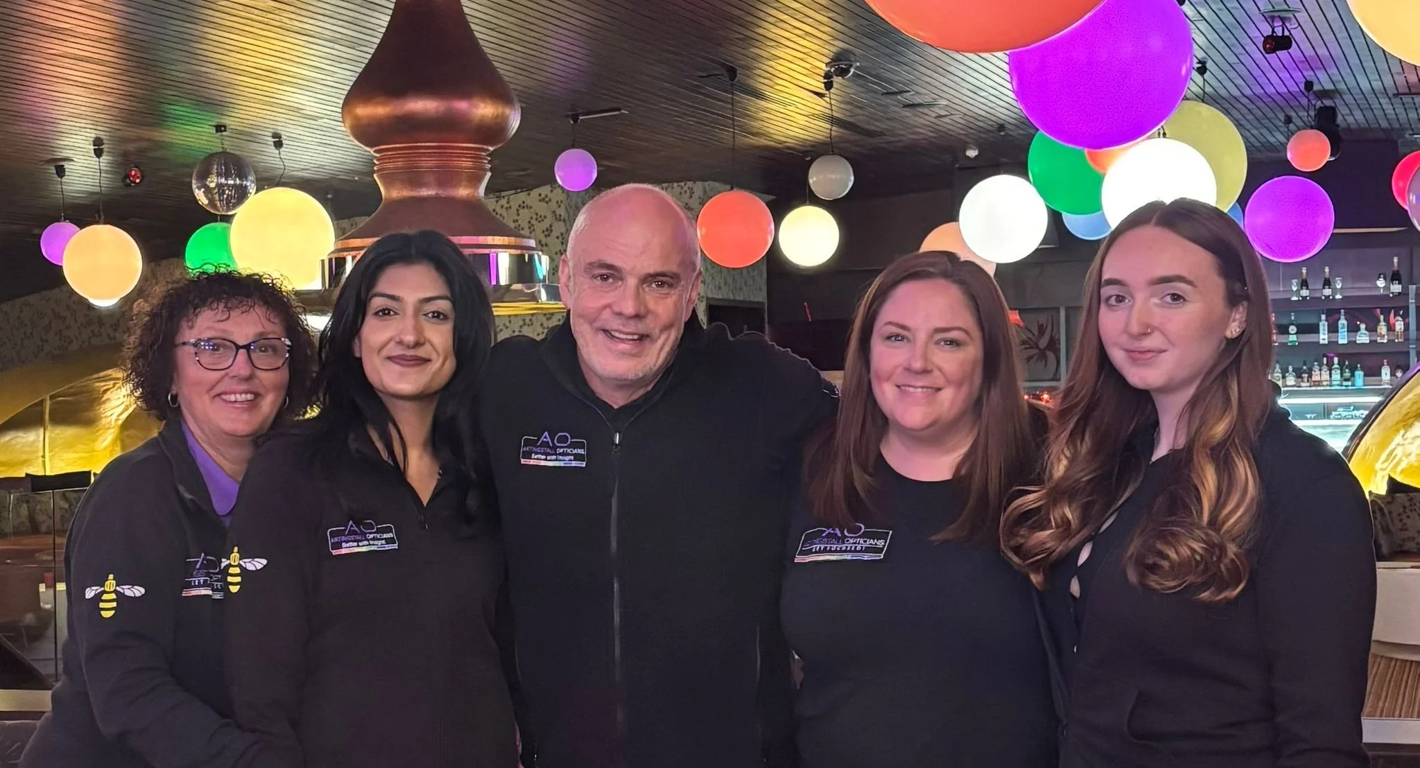 Five people, four women and one man, standing together and smiling inside a restaurant or bar with colorful hanging lights and a bar in the background.
