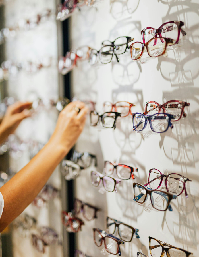 Person shopping for eyeglasses at an eyewear store, browsing a display of various colorful eyeglasses on a wall.