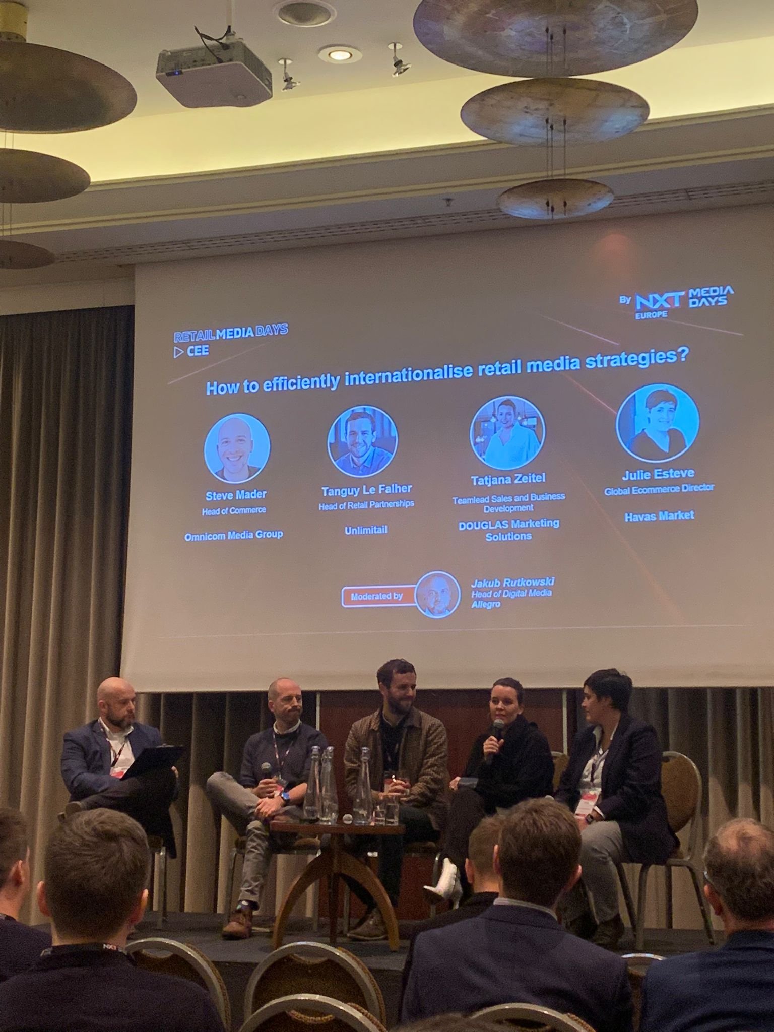 Bühne mit einem Panel von fünf Personen bei einer Konferenz, darunter zwei Frauen und drei Männer, mit einem großen Bildschirm im Hintergrund, der den Titel und die Teilnehmer einer Diskussion über internationale Retail-Medien-Strategien zeigt.