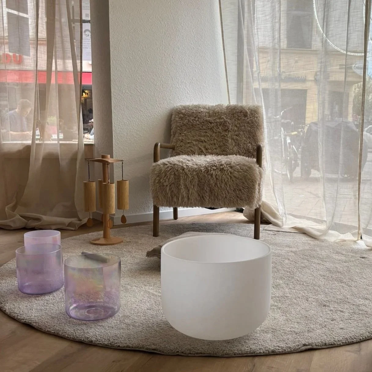 Gemütliches Studio mit einem Sessel mit pelzigem Bezug, einer runden weißen Klangschale, drei lila Kristallklangschalen und einem Steh-Objekt mit Holzlamellen, vor einem Fenster mit transparenten Vorhängen.