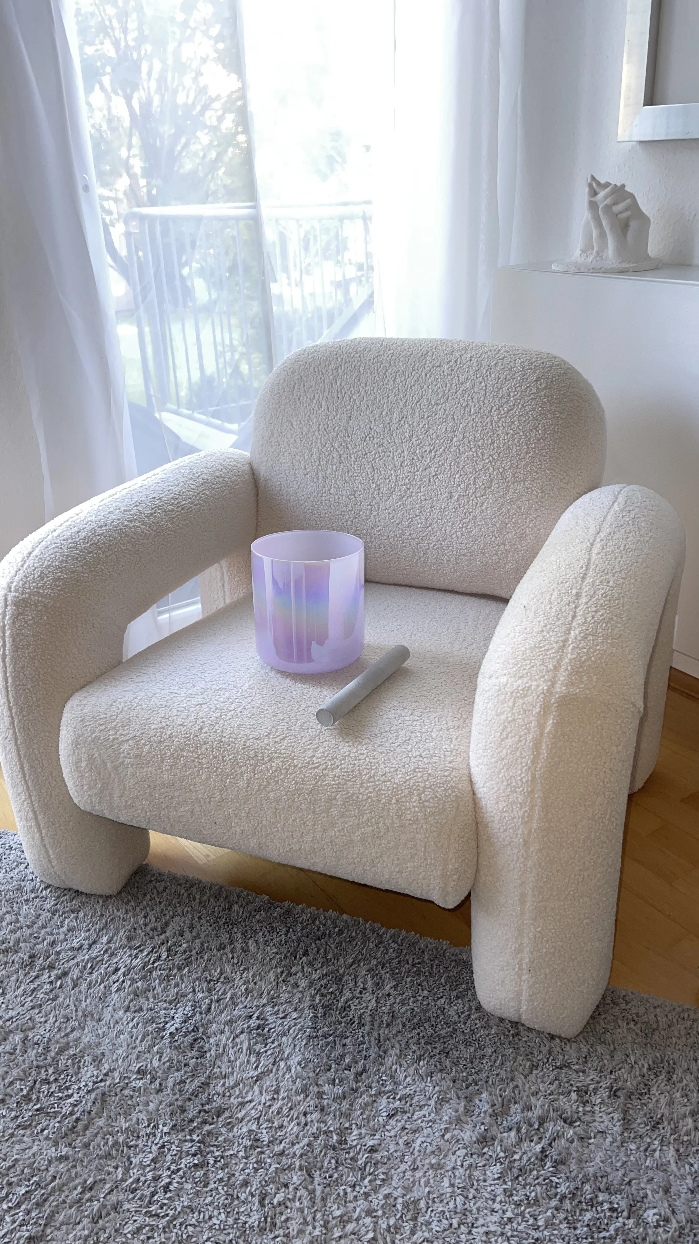 Ein gemütlicher Sessel vor einem Fenster mit weißen Vorhängen, auf dem Sessel stehen eine bunte Kristallklangschale und ein grauer Schlegel.