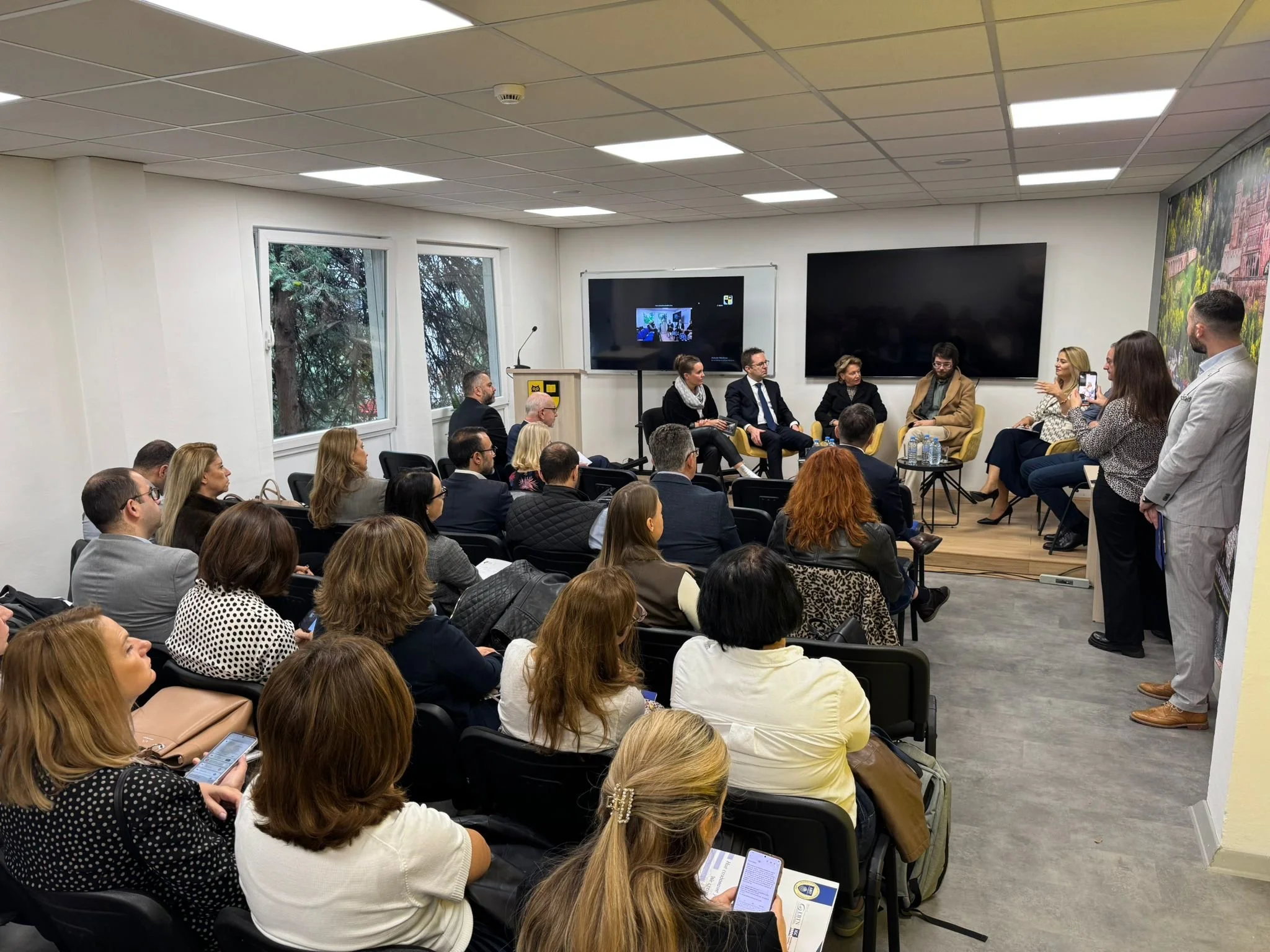 Ein Konferenzraum mit mehreren sitzenden und stehenden Menschen, eine Panel-Diskussion auf der Bühne, zwei große Bildschirme im Hintergrund, einige Leute nehmen Fotos auf.