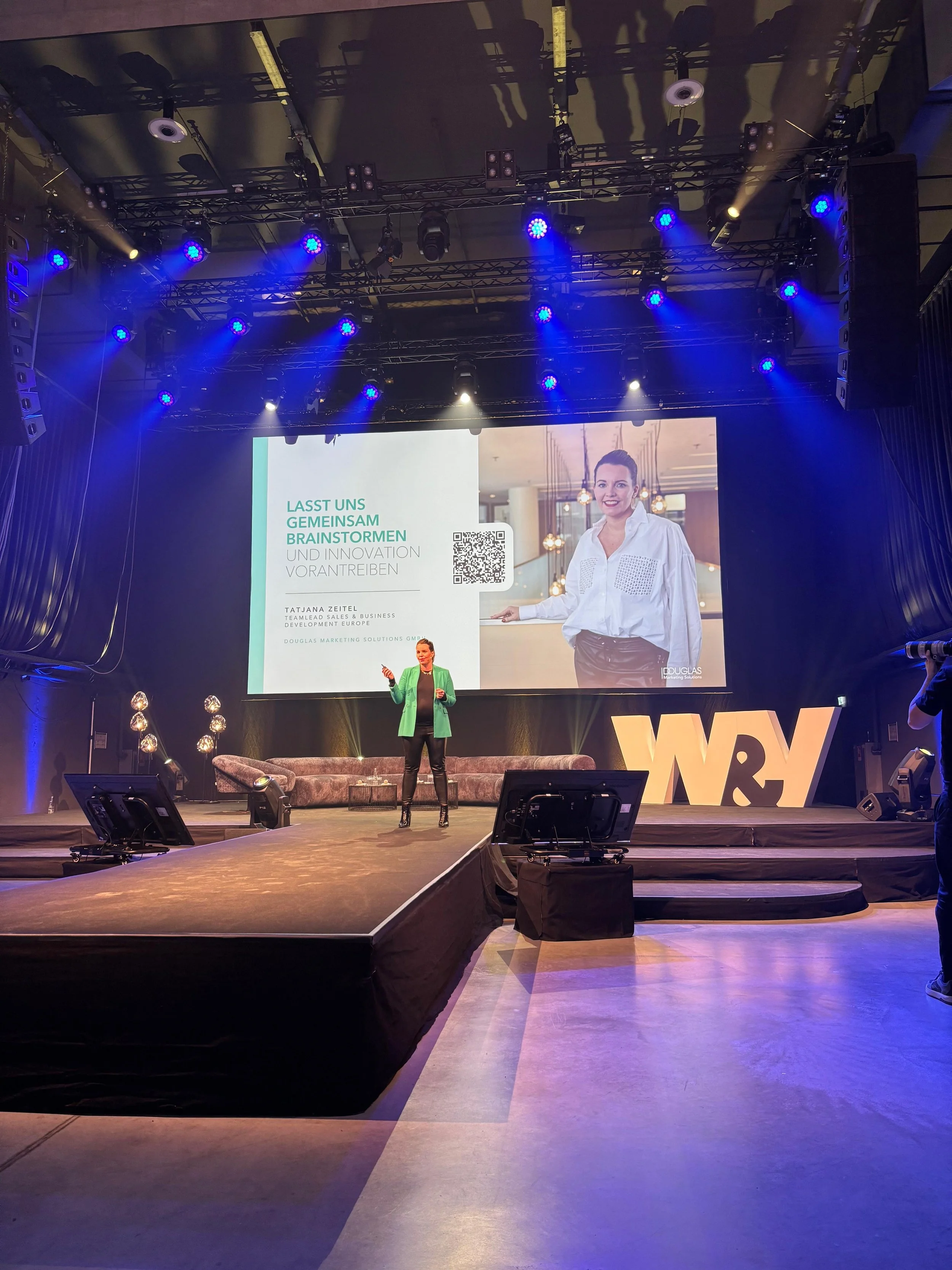Eine Frau hält eine Präsentation auf einer Bühne bei einer Konferenz mit großen Bühnenbild. Im Hintergrund ist eine große Leinwand mit einem Bild einer lächelnden Frau, die im weißen Hemd gekleidet ist. Es sind Scheinwerfer und Lautsprecher zu sehen.