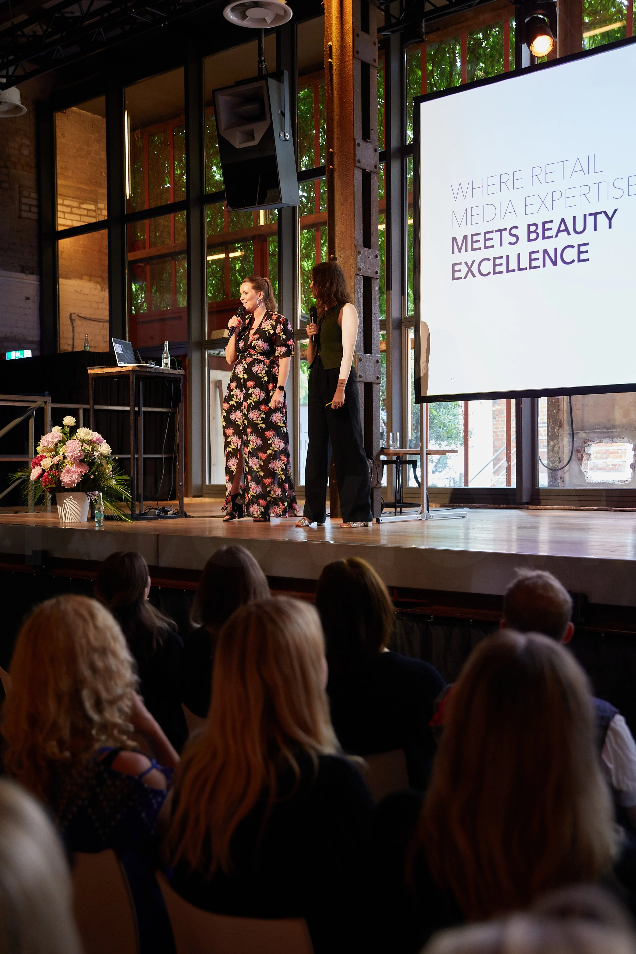Zwei Frauen sprechen auf einer Bühne bei einer Konferenz, im Hintergrund ein Bildschirm mit Text und ein Blumenarrangement auf der linken Seite.