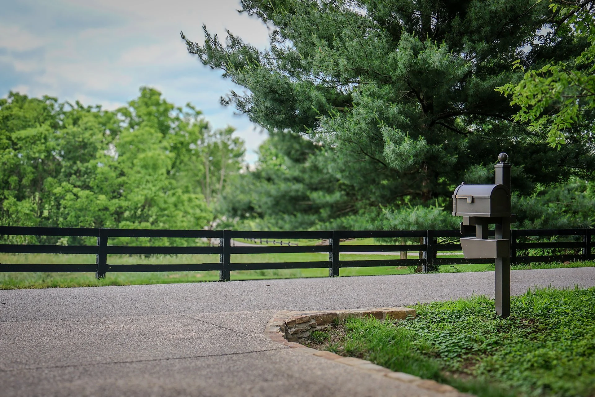A black mailbox on a post next to a paved pathway, with a green grassy area and trees in the background.