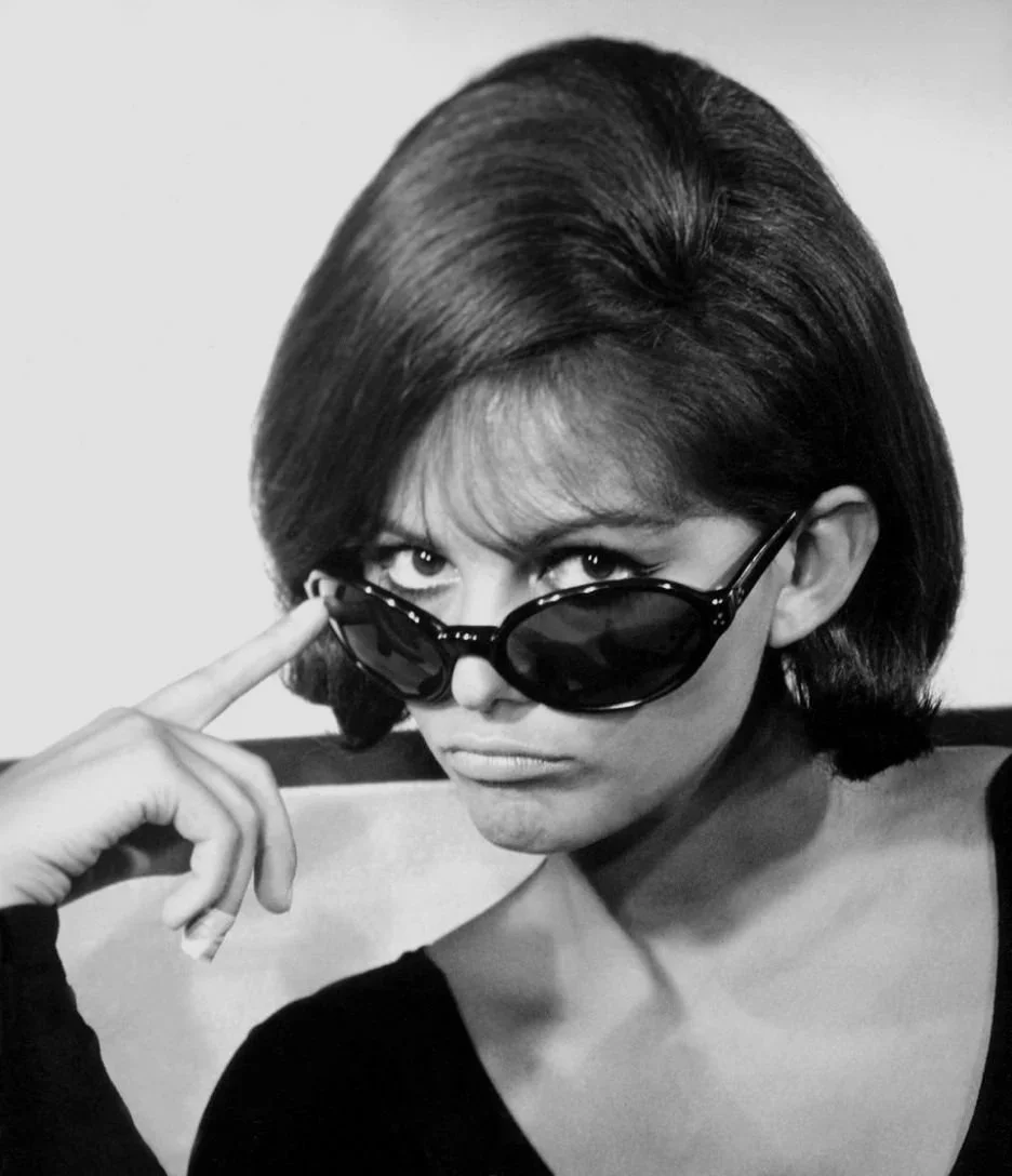 Claudia Cardinal wearing sunglasses, looking over them, with a serious expression.