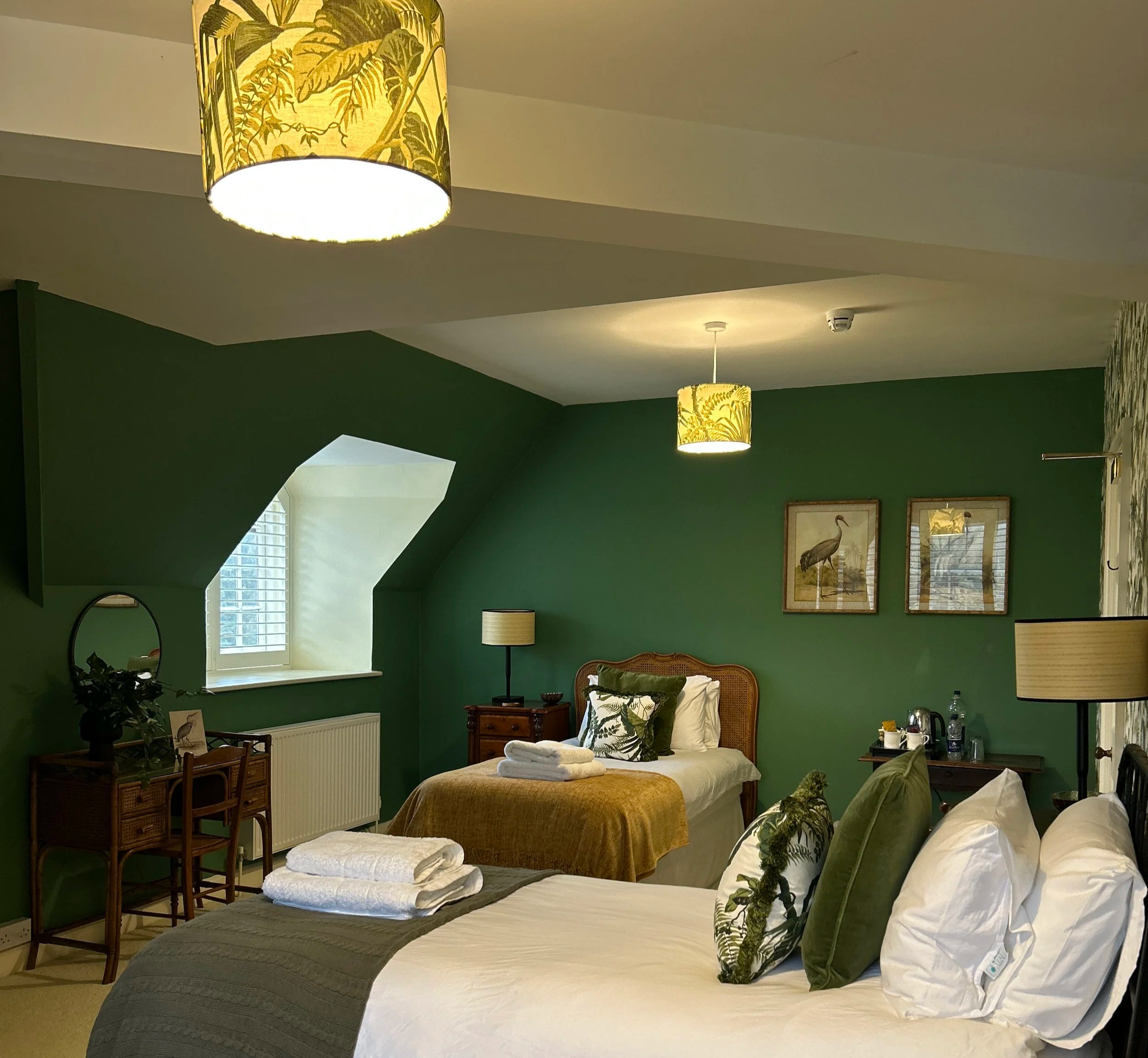 Stunning bedroom at Little Massingham Manor with green feature wall, two beds with white and green pillows, wooden furniture, framed bird artwork, and hanging yellow lampshades with botanical designs.