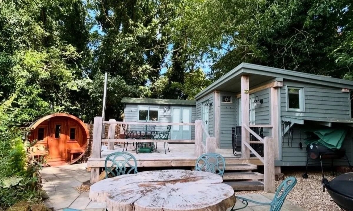Beautiful Woodland Lodge in the grounds of Little Massingham Manor with a wooden deck, outdoor furniture, a clay cooking oven and surrounded by green trees and foliage.