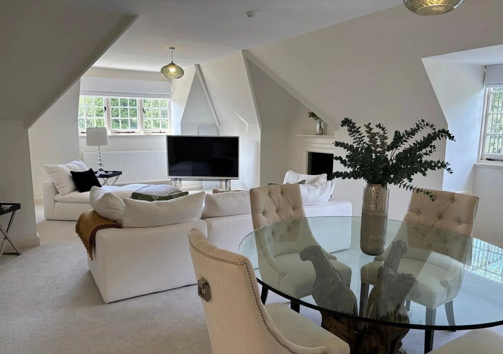 Living room with white sofas, a glass dining table with cream chairs, a large vase with green foliage, windows letting in natural light, a black television, fireplace, and a side table with lamp.