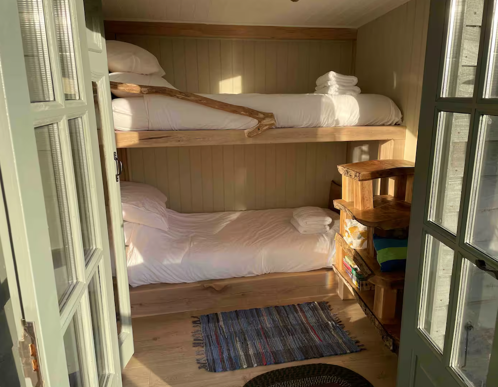 A cozy room with a wooden bunk bed, white bedding on both beds, and towels folded on the top bunk. There is a small wooden bookshelf with books and a blanket, a colorful striped rug on a wooden floor, and double glass doors that open to the room.