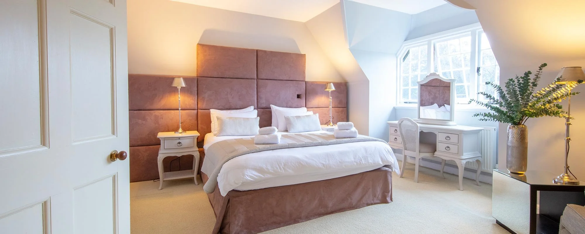 A cozy bedroom with a large bed, pink padded headboard, white bedding, and neatly folded towels on top. There are two bedside tables with lamps, a white dressing table with a mirror, and a mirror chair near a window. A tall potted plant and a lamp ar
