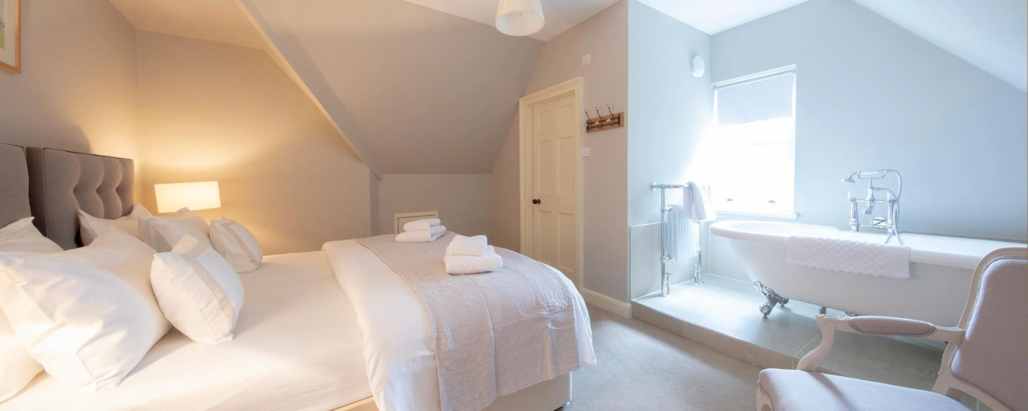 A bright and cozy bedroom with a large bed, white bedding, and multiple pillows, featuring a lamp on the side table. Adjacent to the bed is a bathroom area with a vintage-style bathtub, a towel hanging over it, and a chair nearby, illuminated by a wi