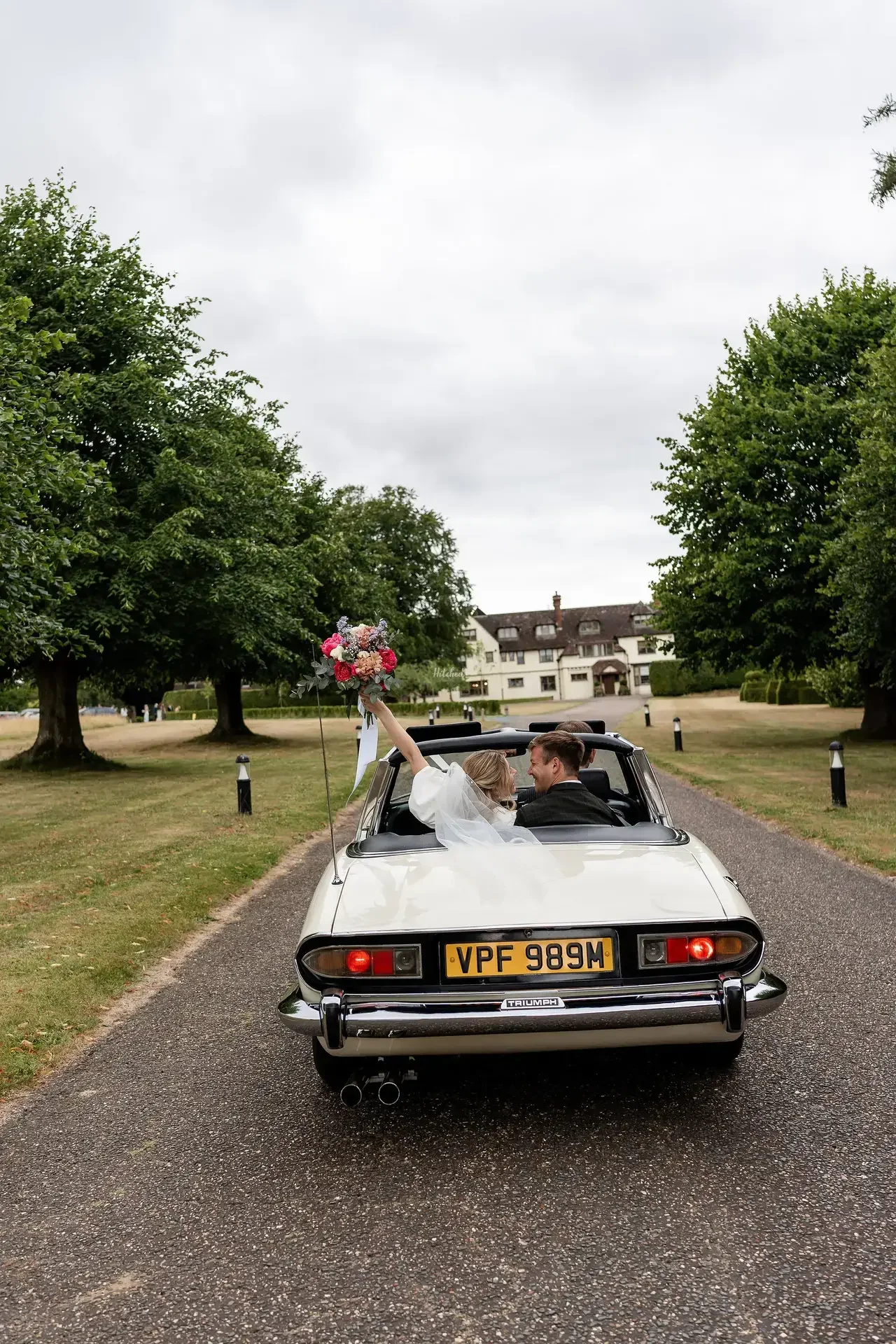 A bride and groom sitting in a vintage white convertible car on a driveway heading to Little Massingham Manor, with the bride holding a colourful bouquet in the air, celebrating a wedding outdoors on a cloudy day.