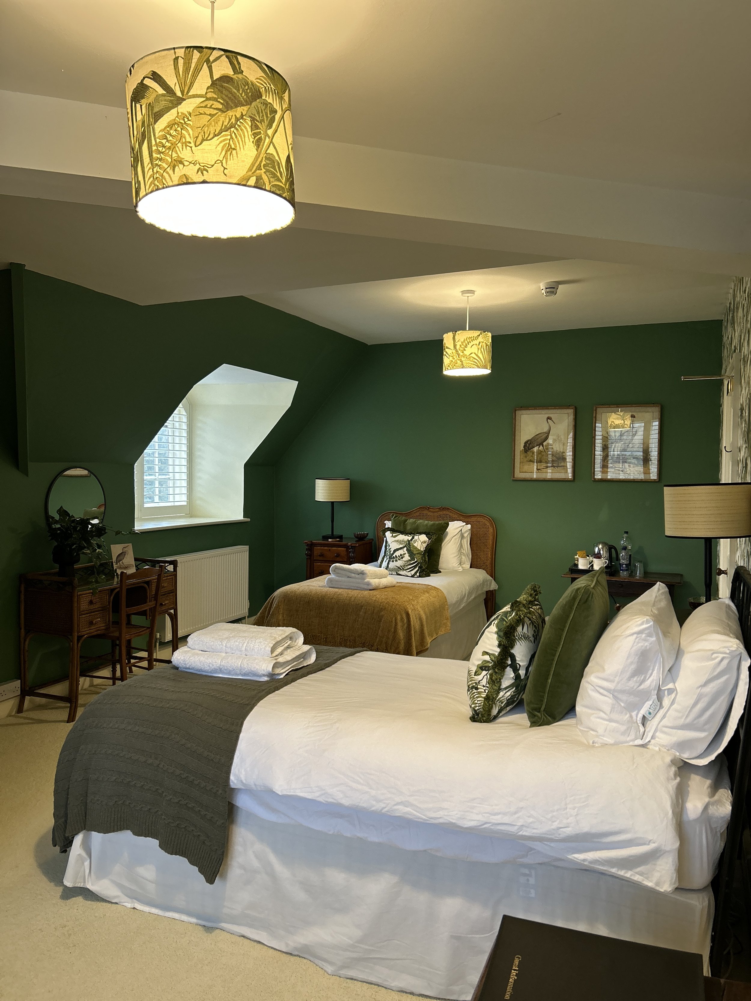 Stunning bedroom at Little Massingham Manor with green feature wall, two beds with white and green pillows, wooden furniture, framed bird artwork, and hanging yellow lampshades with botanical designs.