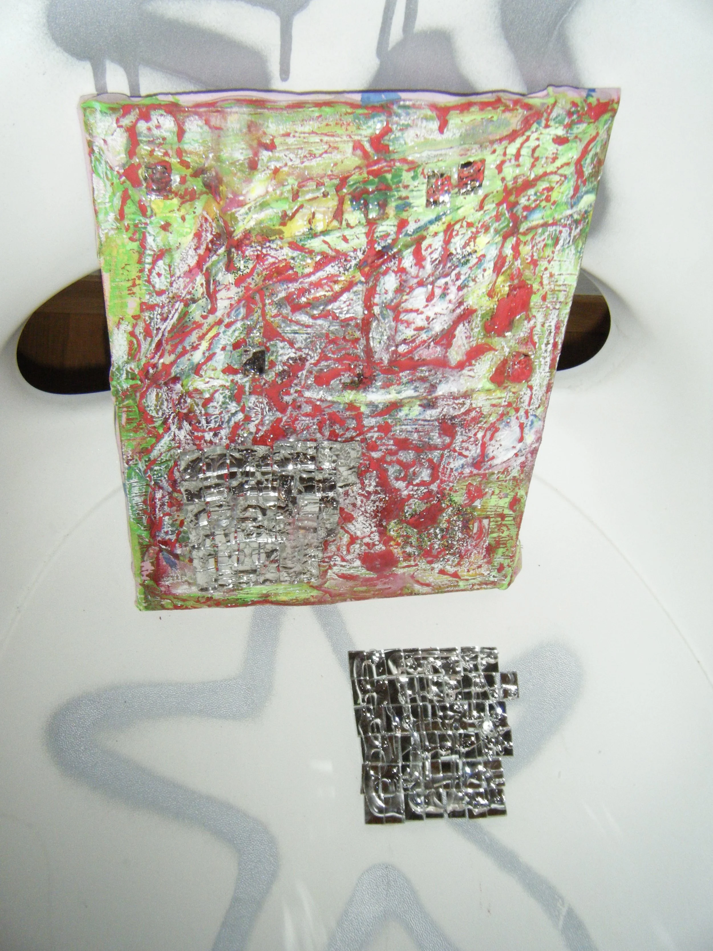 An abstract mixed media art piece with vibrant splashes of pink, green, yellow, and red, including metallic silver elements, placed on a white table with a gray patterned tablecloth underneath. A separate metallic silver grid component is also on the table.