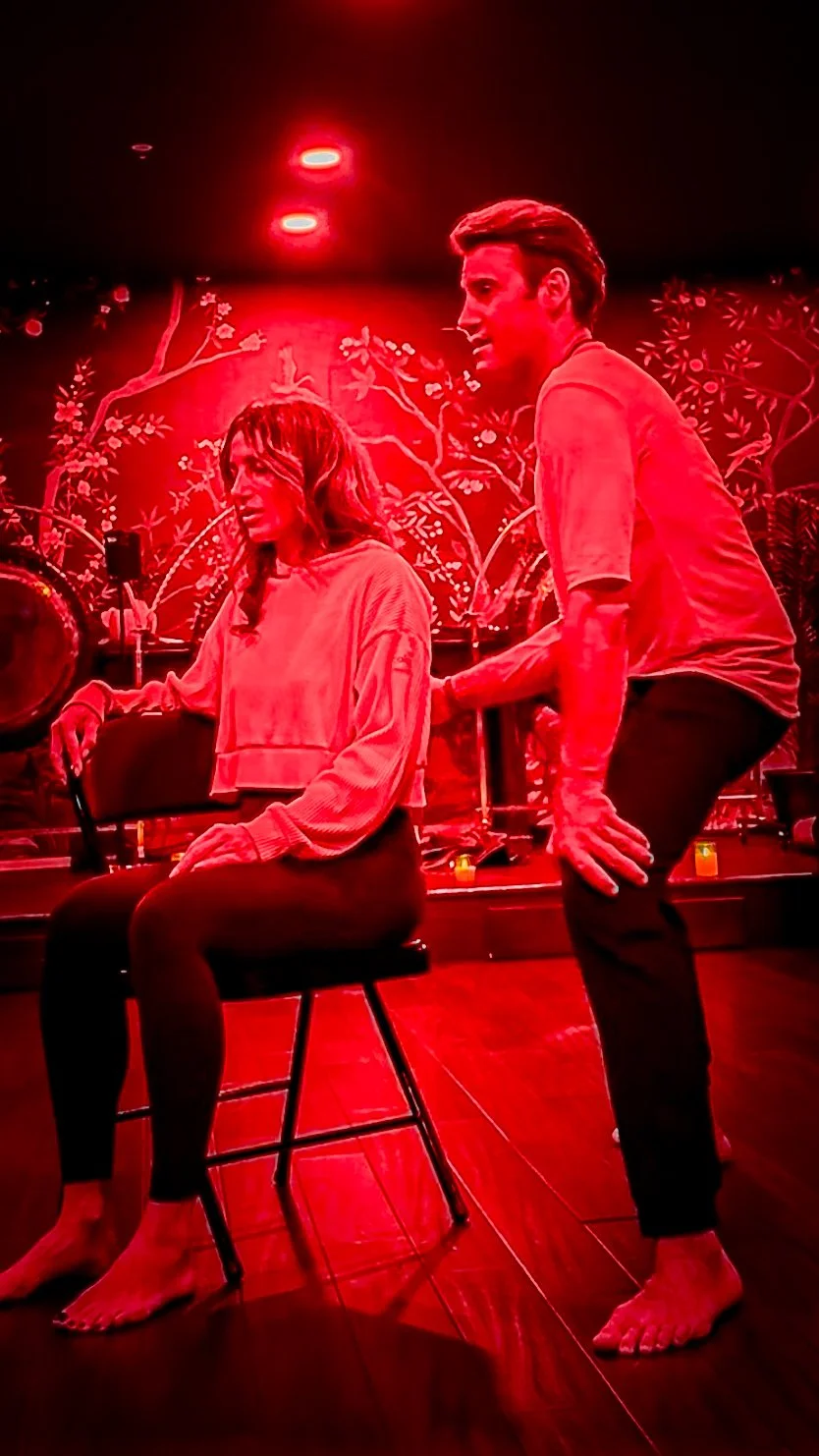 A man and a woman in a dimly lit room with vibrant red lighting and a decorative mural backdrop. The woman is sitting on a chair, holding a microphone, while the man is squatting beside her, touching her arm and engaging in conversation. Both are barefoot.